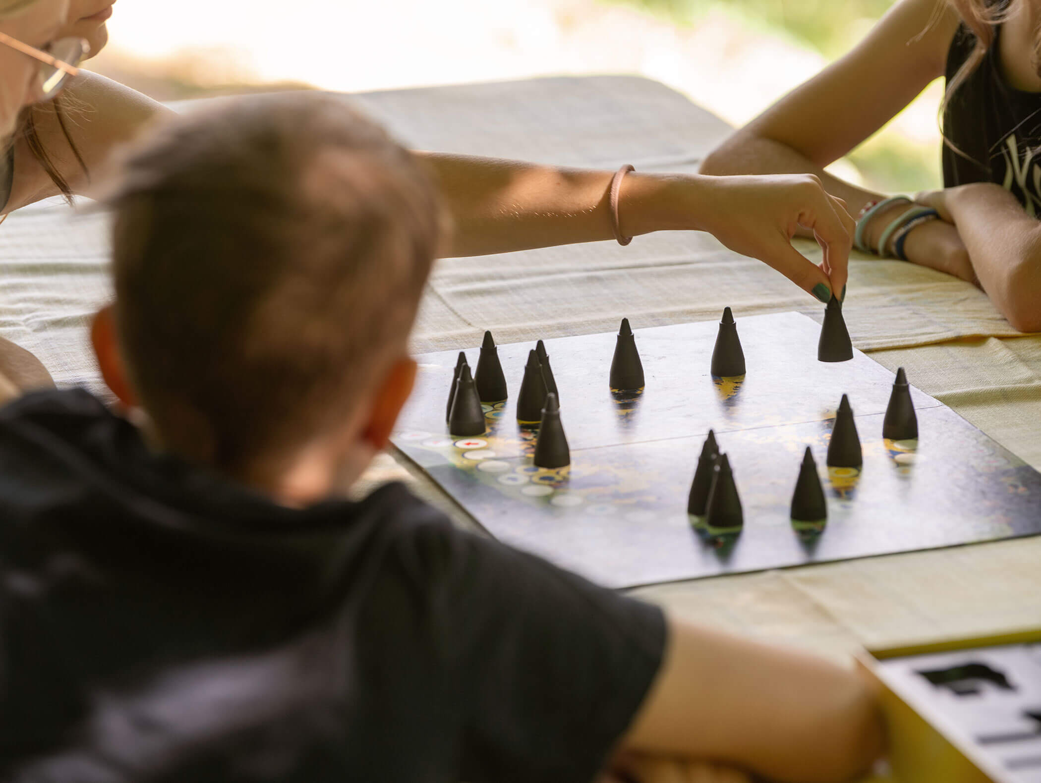 Drei Personen spielen ein Brettspiel mit schwarzen kegelförmigen Spielsteinen auf einem mit einem hellen Tuch bedeckten Tisch. - Wandergasthof Steinegger