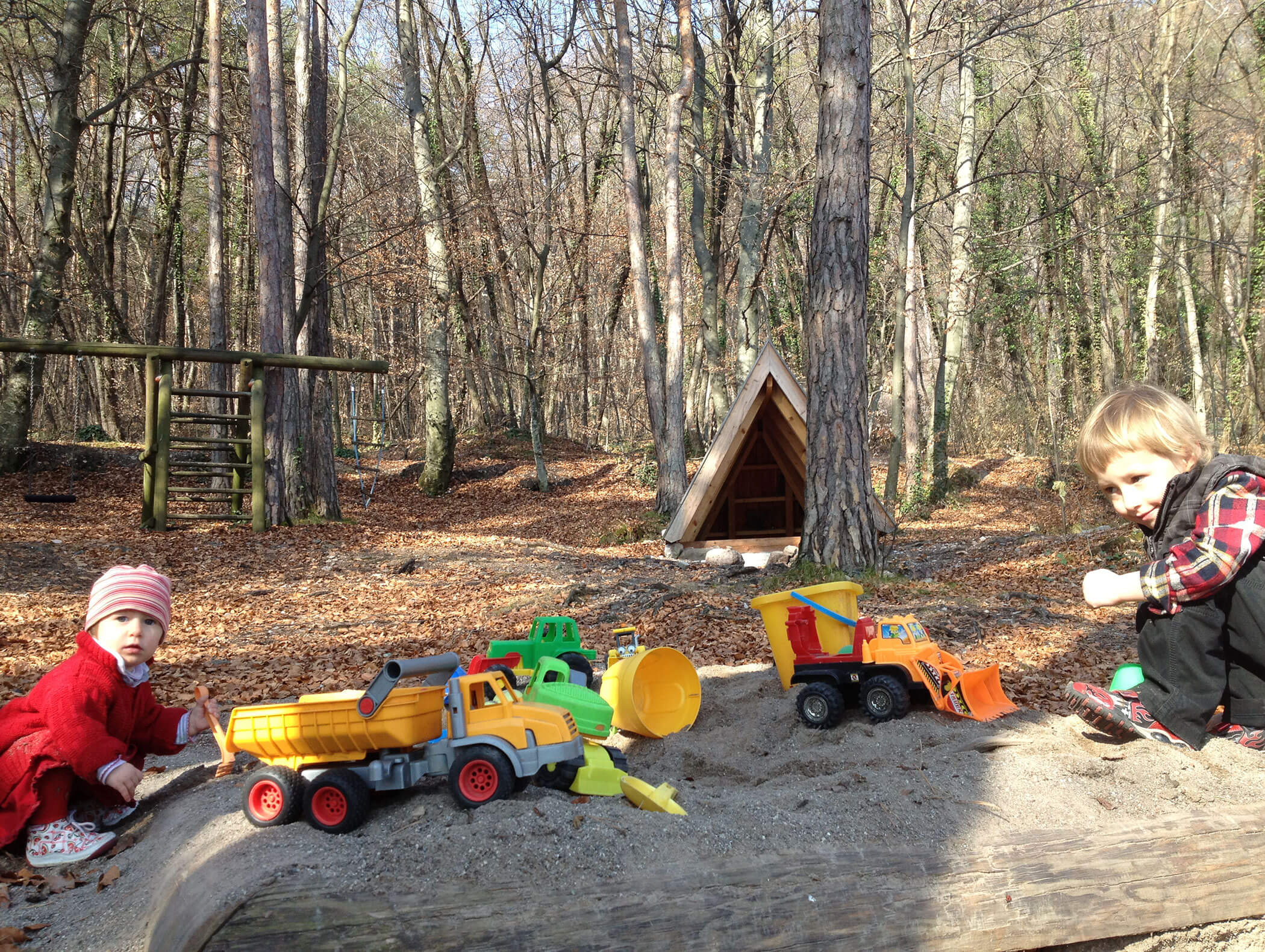 Zwei Kinder spielen mit Spielzeugautos auf einer sandigen Fläche in einem Wald neben einer kleinen Holzhütte. - Wandergasthof Steinegger