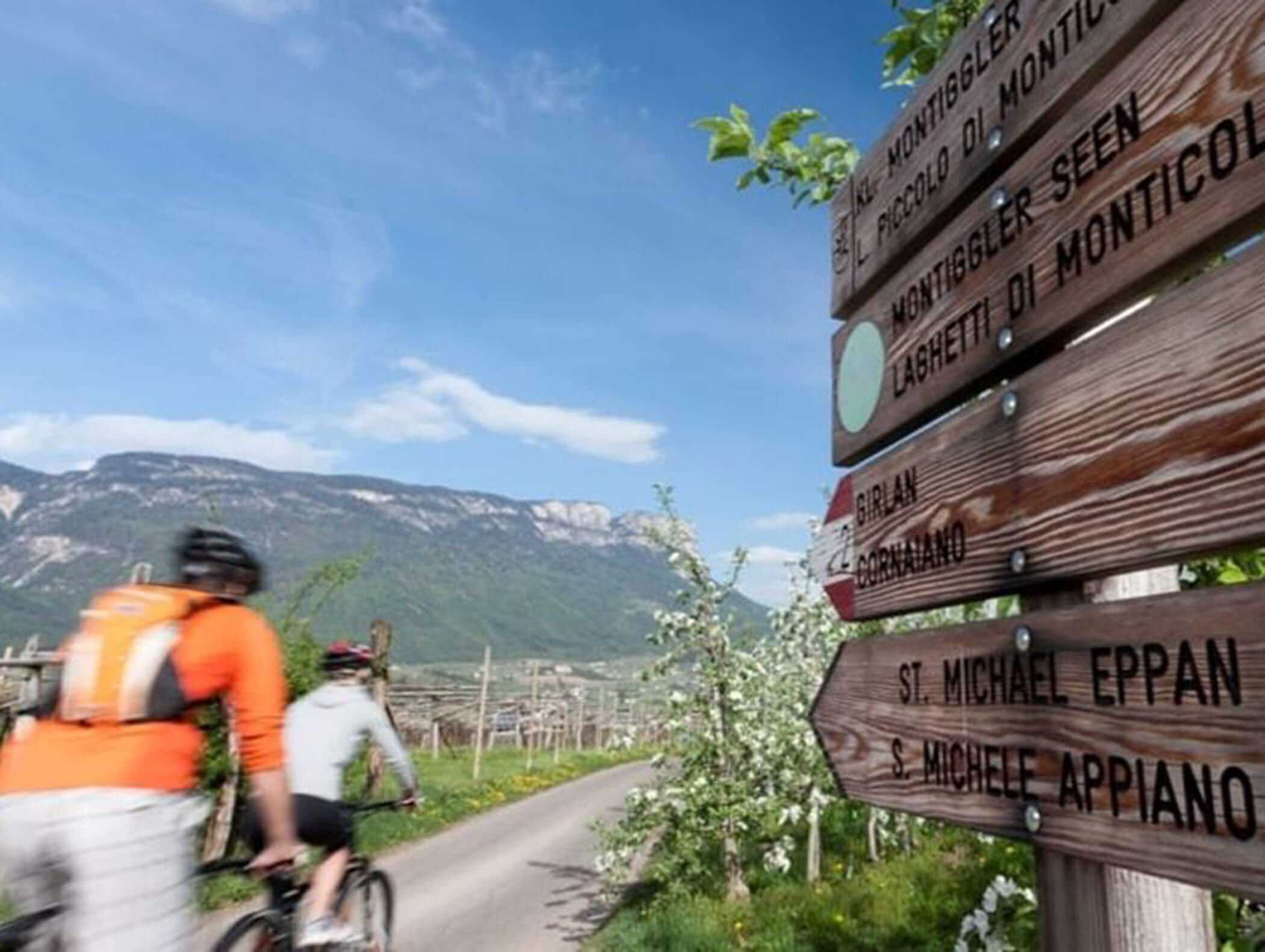 Zwei Radfahrer fahren auf einem ländlichen Weg an hölzernen Wegweisern vorbei, mit Bergen und blauem Himmel im Hintergrund. - Wandergasthof Steinegger