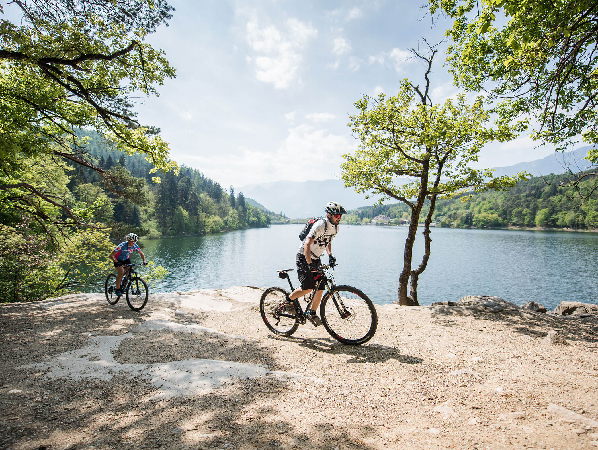 Zwei Personen fahren mit dem Mountainbike auf einem Weg neben einem See, umgeben von Bäumen und Bergen. - Wandergasthof Steinegger