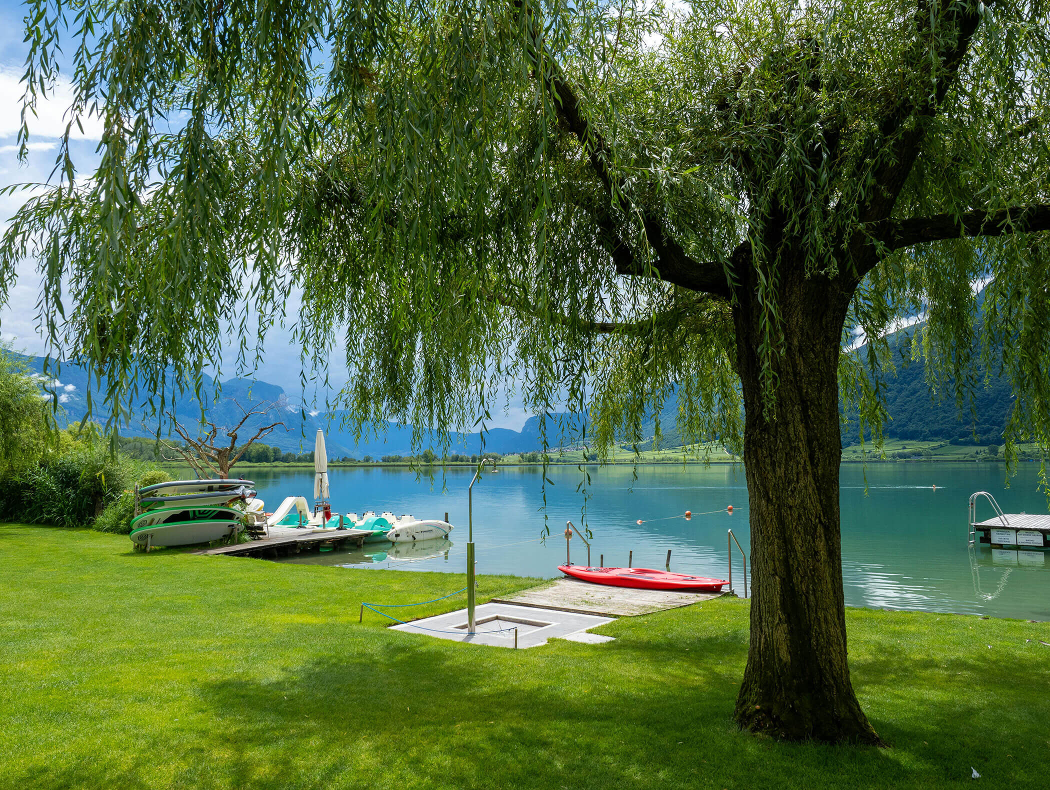 Ein Baum beschattet einen Steg am See mit Booten auf ruhigem Wasser, grünem Gras und Bergen im Hintergrund. - Wandergasthof Steinegger