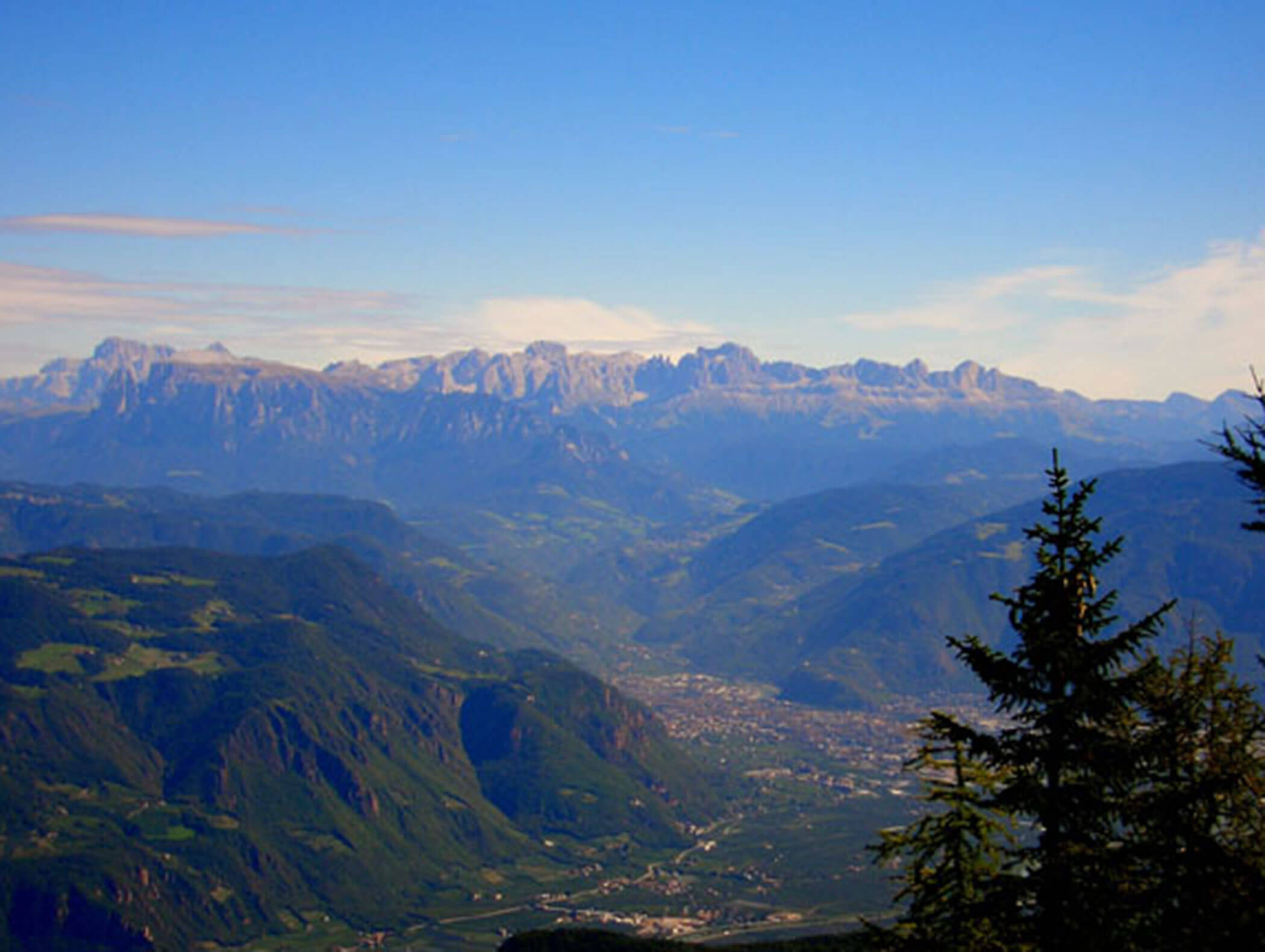 Bergtallandschaft mit fernen Gipfeln, grünen Hügeln und einer kleinen Stadt unter strahlend blauem Himmel. - Wandergasthof Steinegger