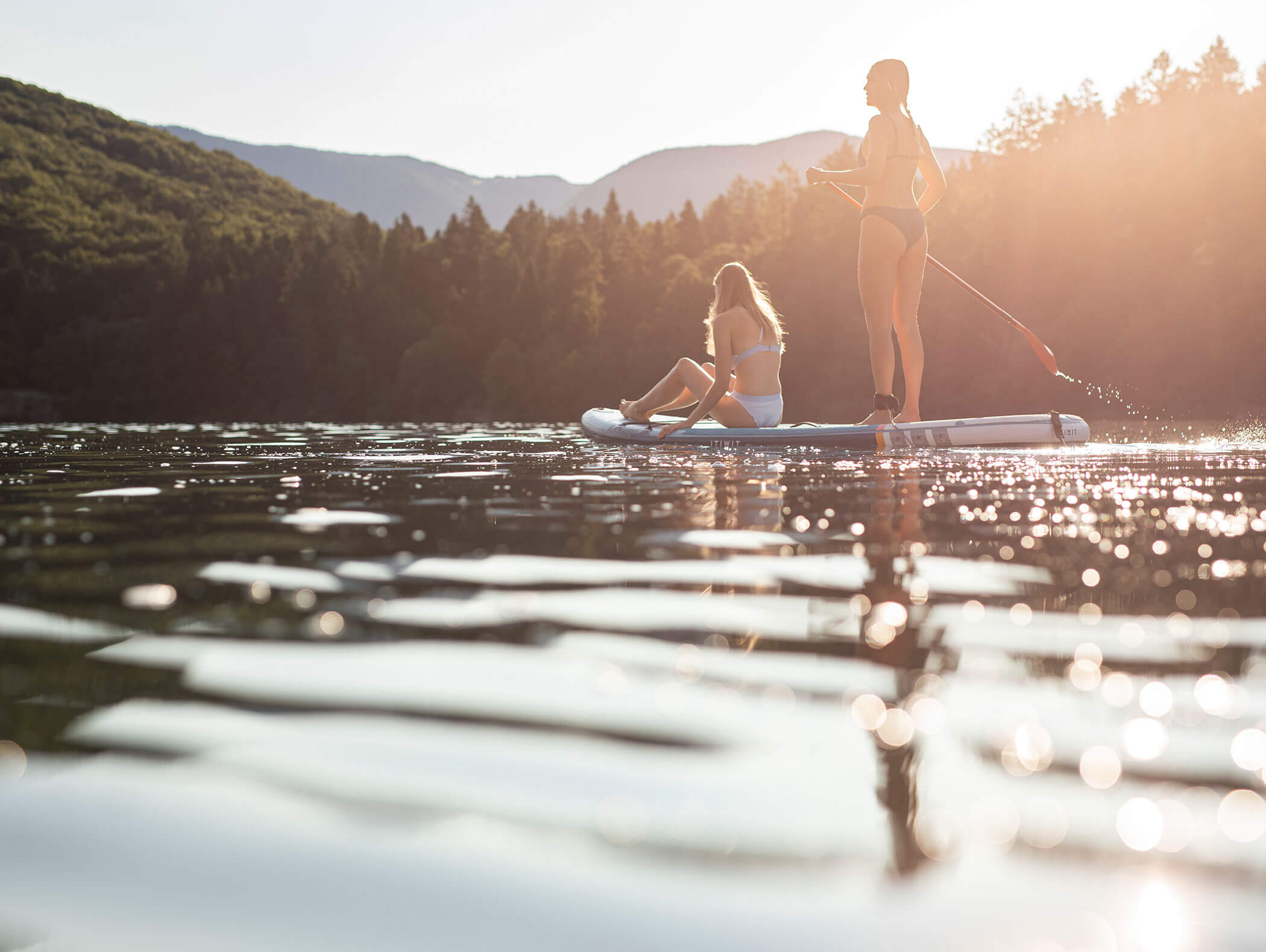 Zwei Frauen beim Paddeln auf einem ruhigen See bei Sonnenuntergang, umgeben von Bäumen und Bergen. - Wandergasthof Steinegger