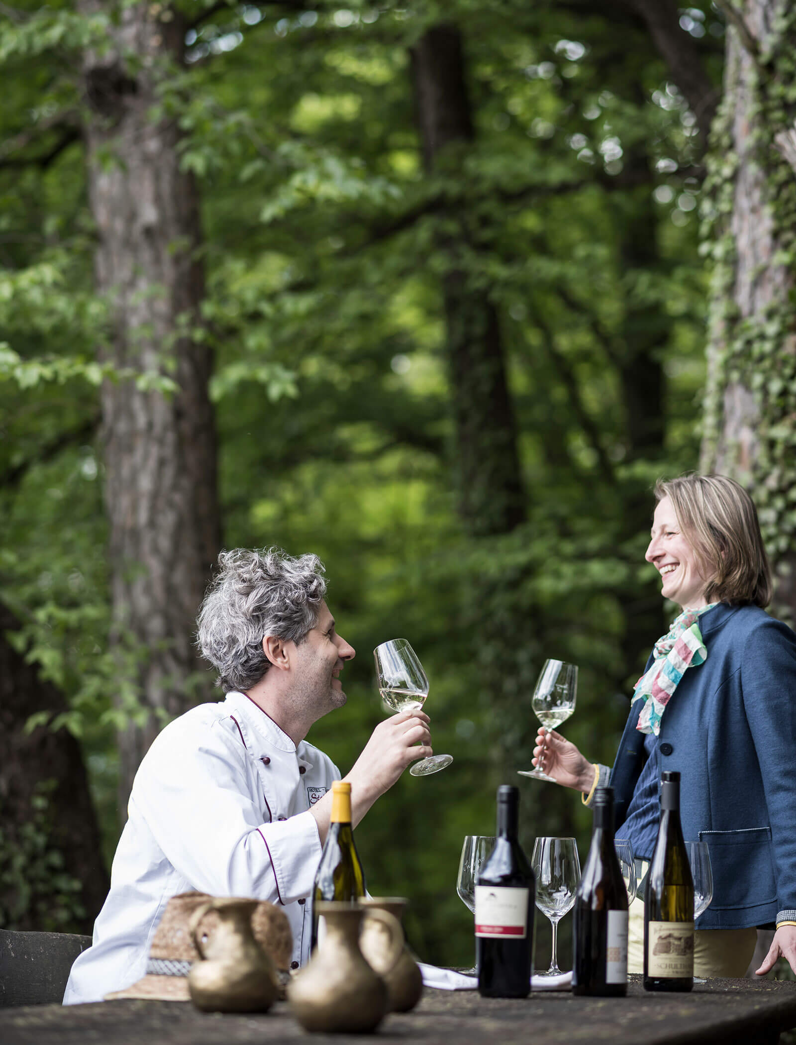 A man and woman clink wine glasses at a table outdoors, surrounded by trees and wine bottles. - Wandergasthof Steinegger