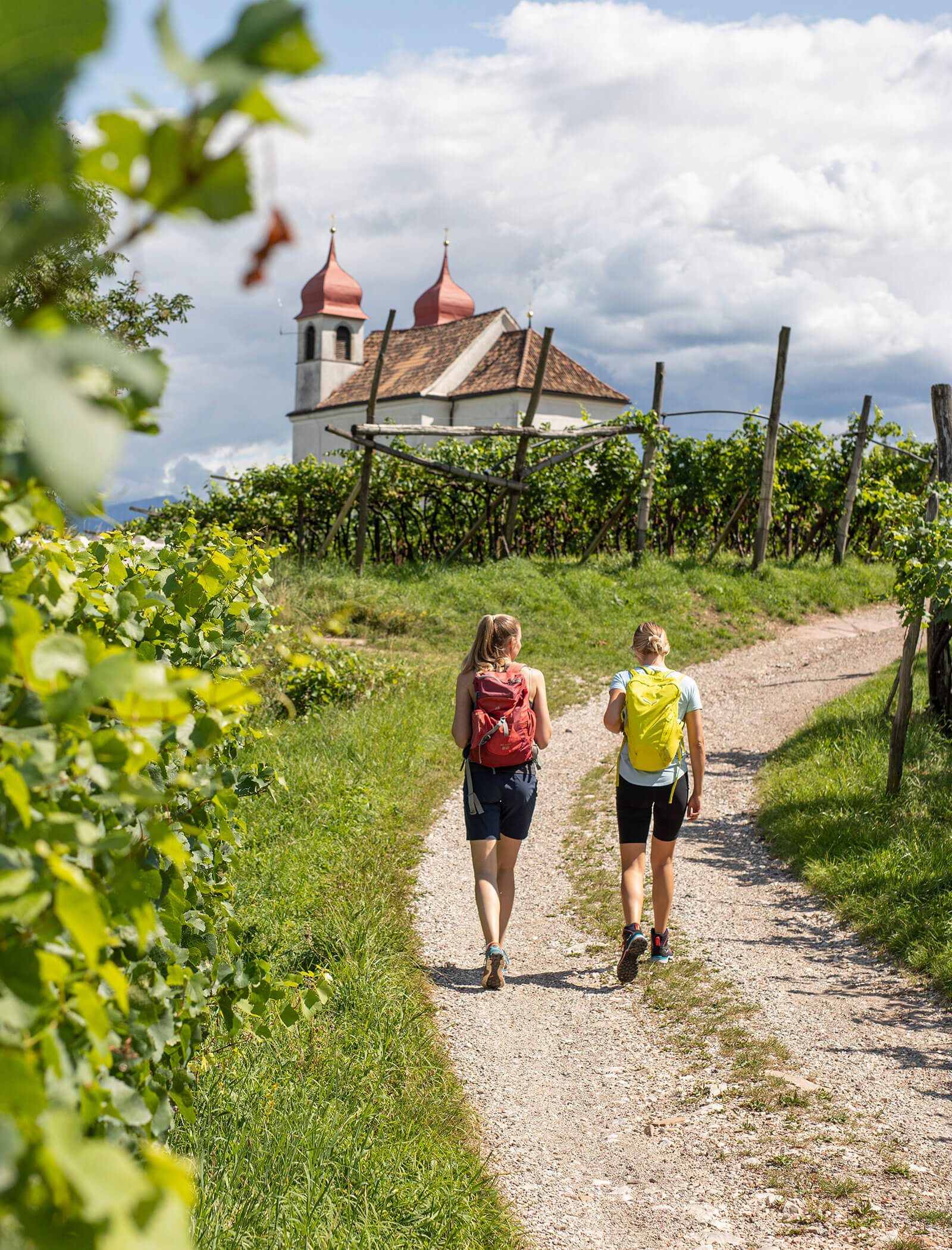 Zwei Frauen wandern auf einem Weinbergsweg in Richtung einer Kirche mit roten Kuppeln unter einem teilweise bewölkten Himmel. - Wandergasthof Steinegger