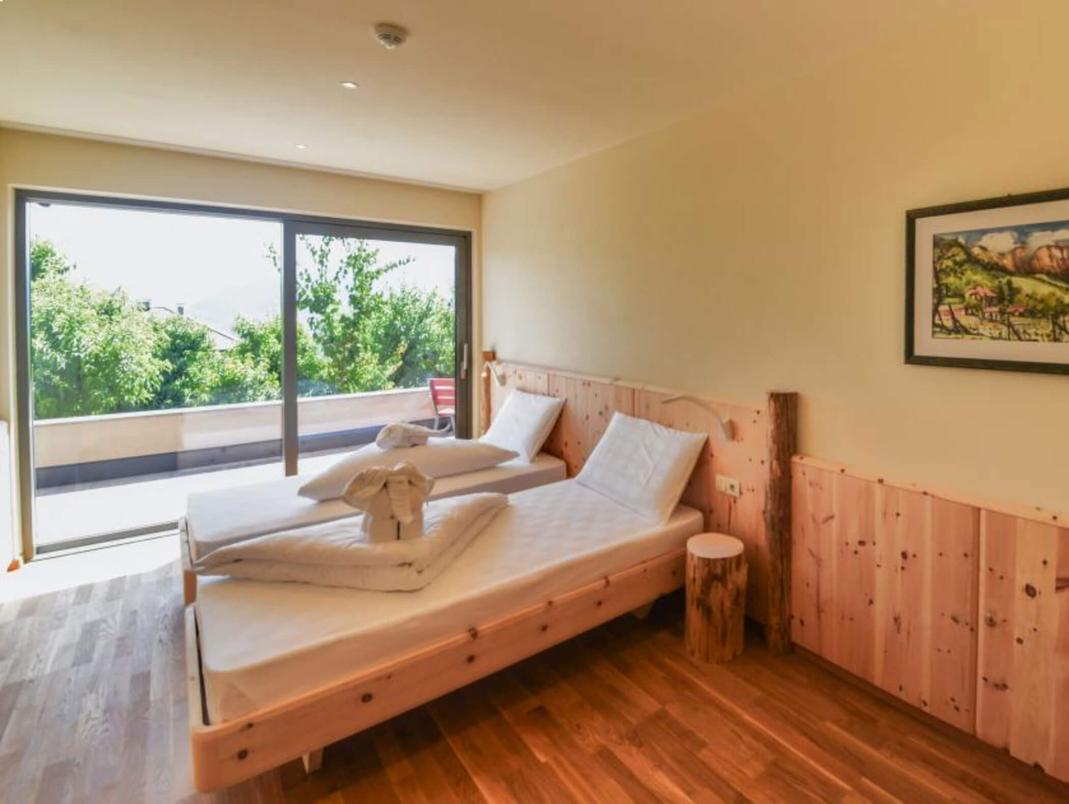 A bright bedroom with wooden furniture, large window, and towel art on the bed overlooking greenery. - Wandergasthof Steinegger