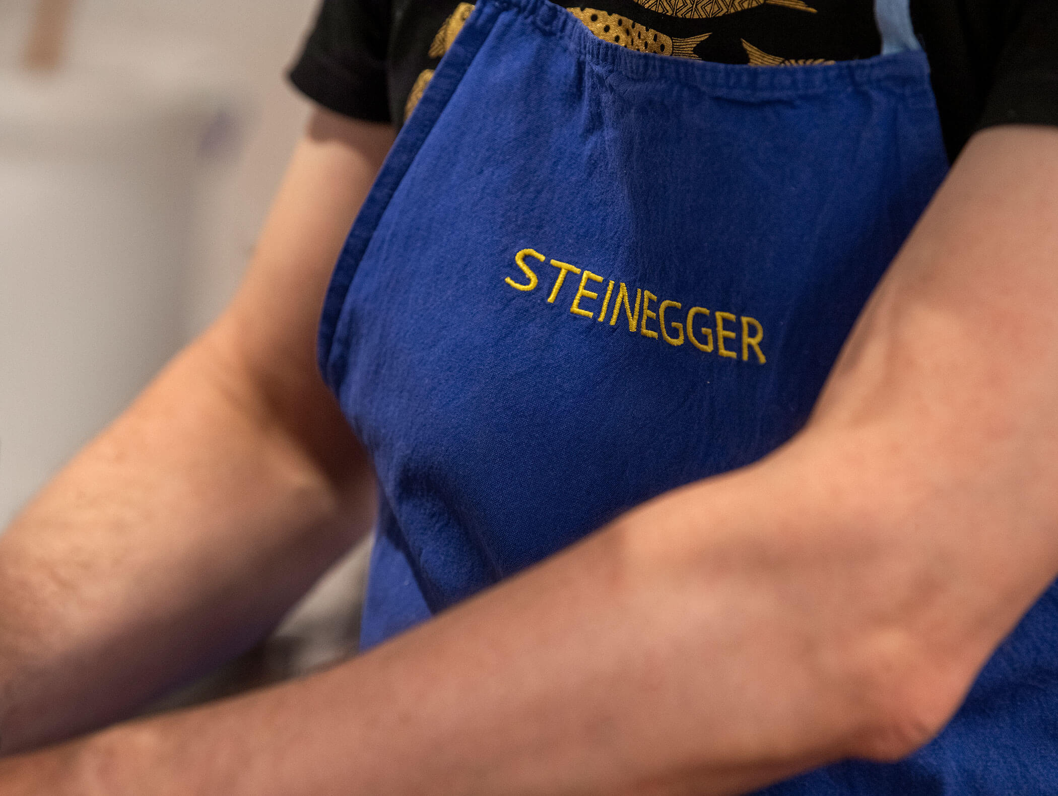 Person, die eine blaue Schürze trägt, auf der in gelben Buchstaben der Name STEINEGGER gestickt ist. - Wandergasthof Steinegger