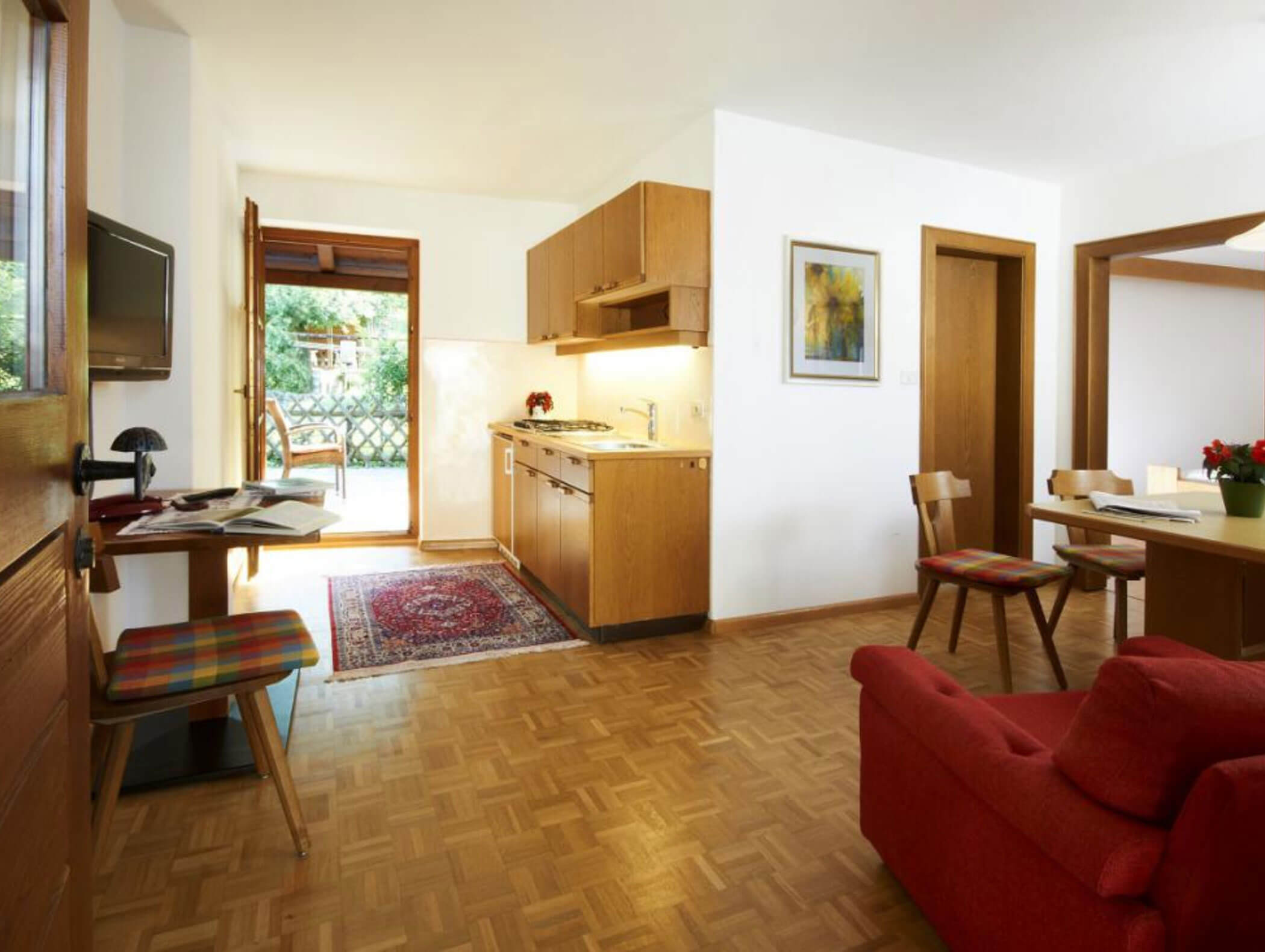 Bright kitchen and living area with wood floors, red sofa, dining table, and open door to a balcony. - Wandergasthof Steinegger