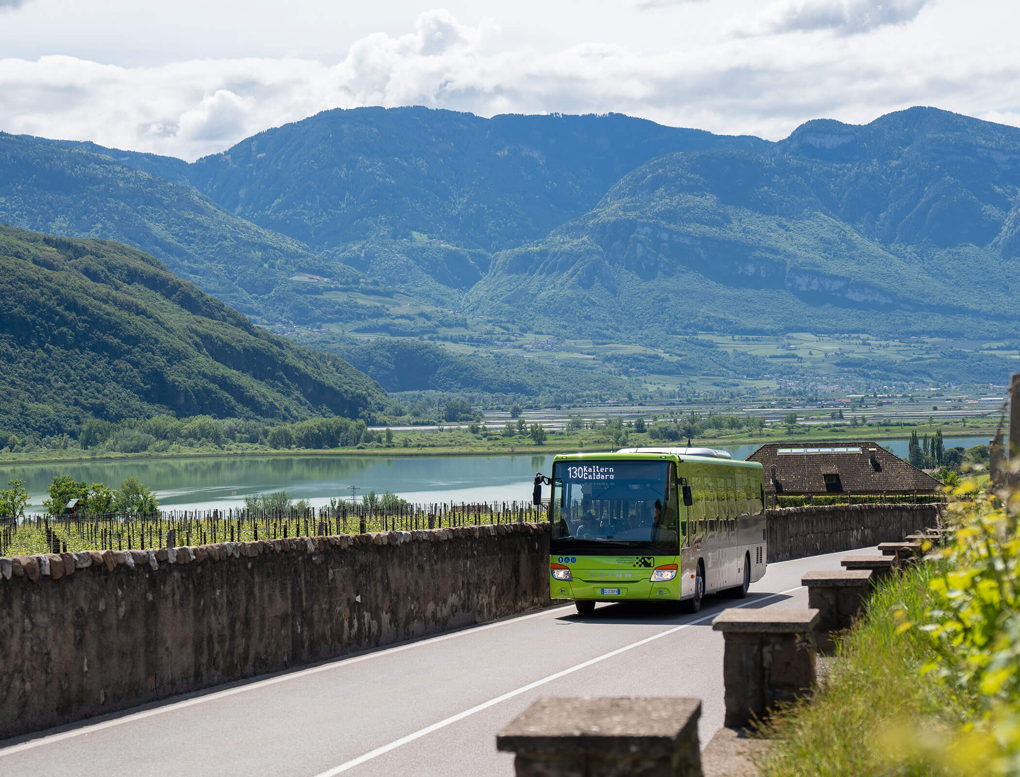 A green bus drives on a road by a lake, with mountains and greenery in the background under a partly cloudy sky. - Wandergasthof Steinegger