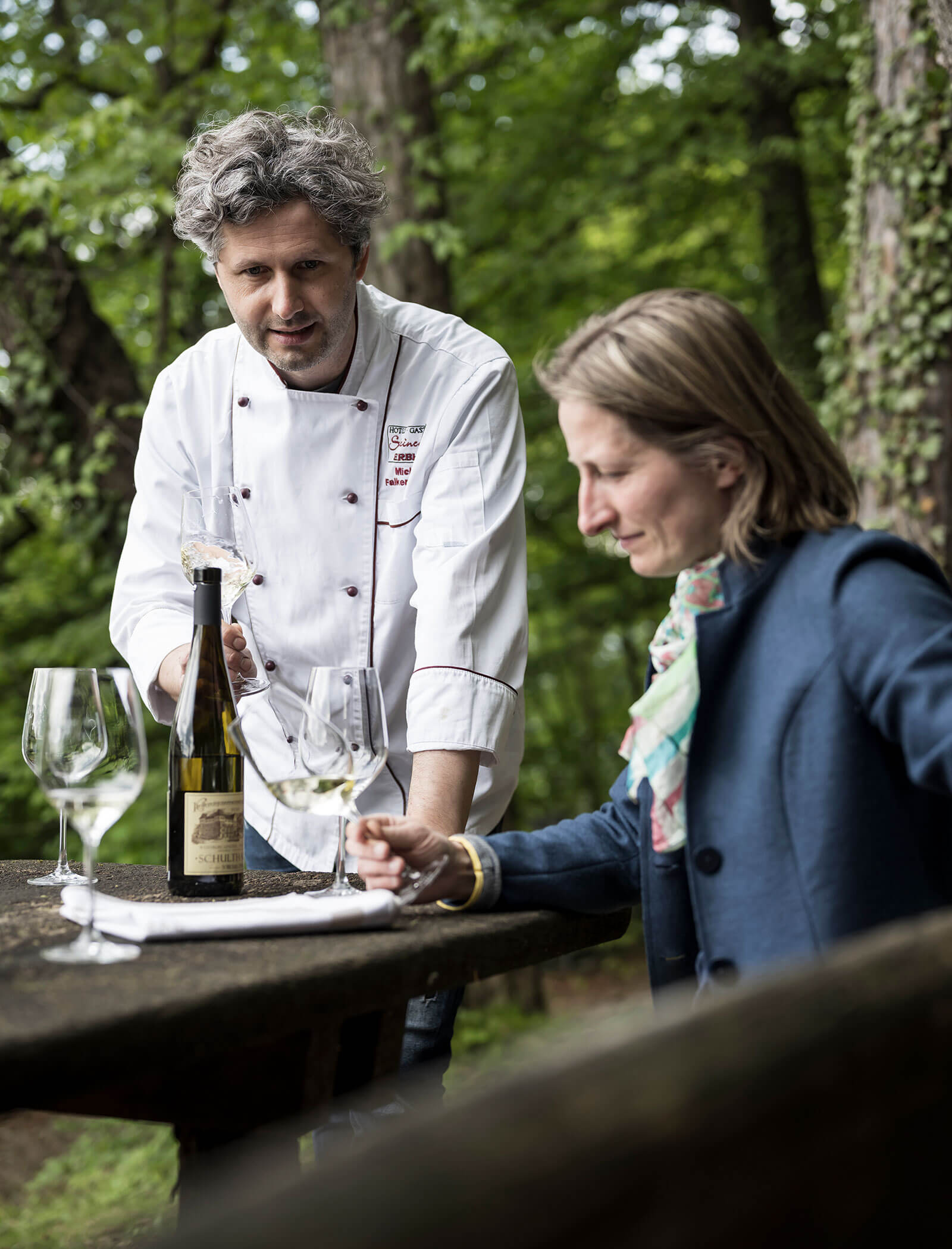 A chef pours white wine for a seated woman at an outdoor table surrounded by trees. - Wandergasthof Steinegger