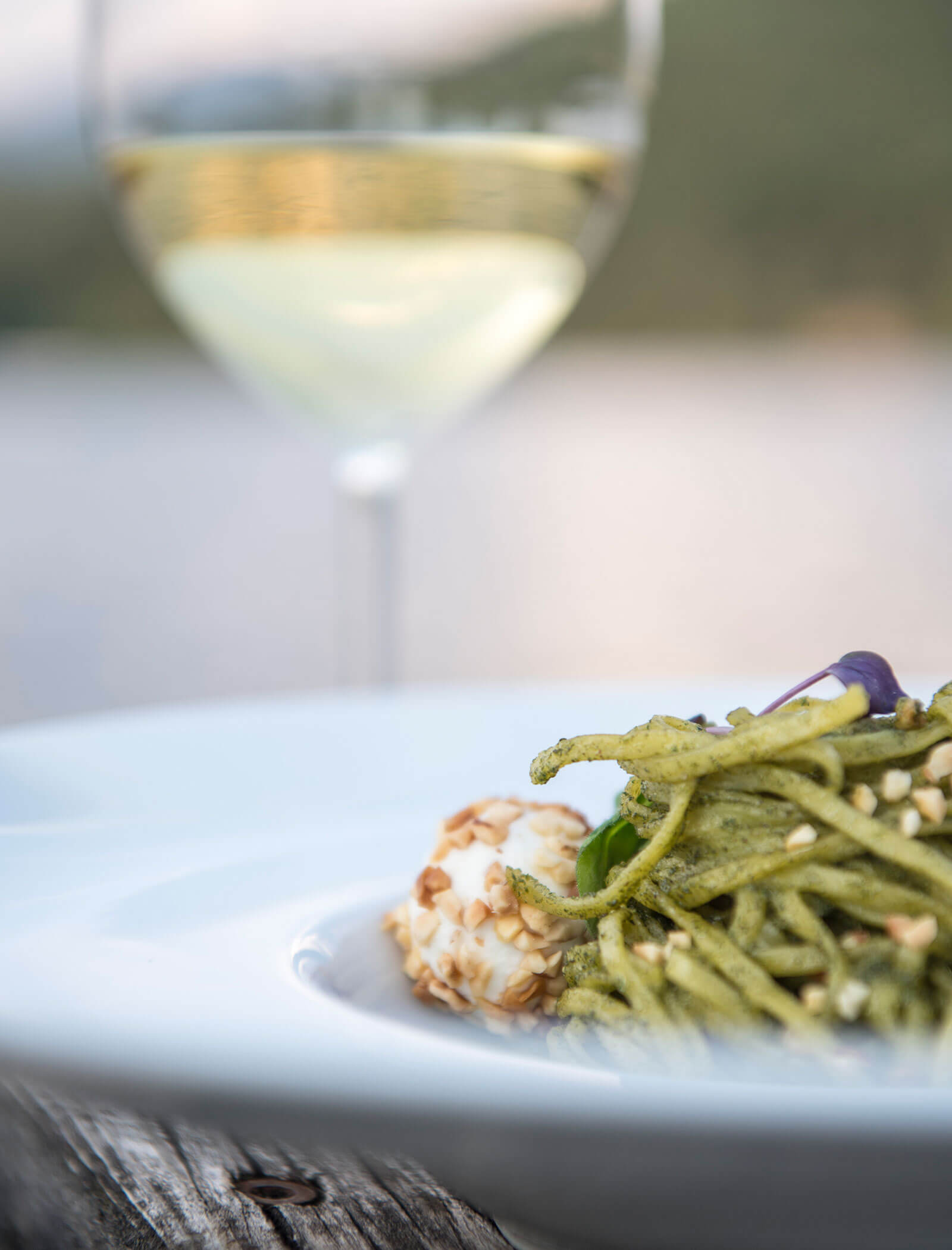 A plate of pesto pasta with cheese, beside a glass of white wine, outdoors. - Wandergasthof Steinegger