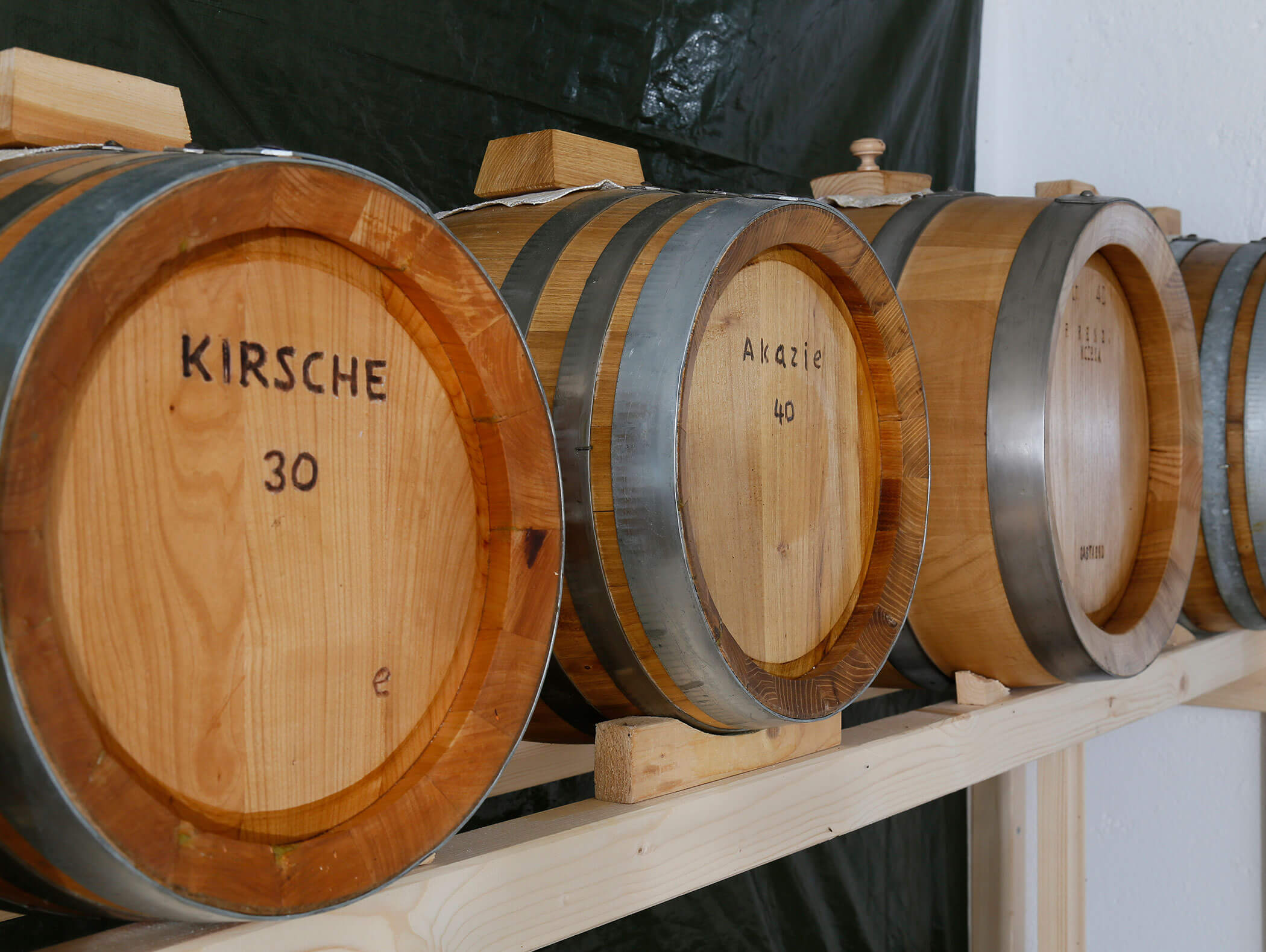 Barili di legno etichettati Kirsche e Akazie si trovano su uno scaffale di legno su uno sfondo scuro. - Wandergasthof Steinegger