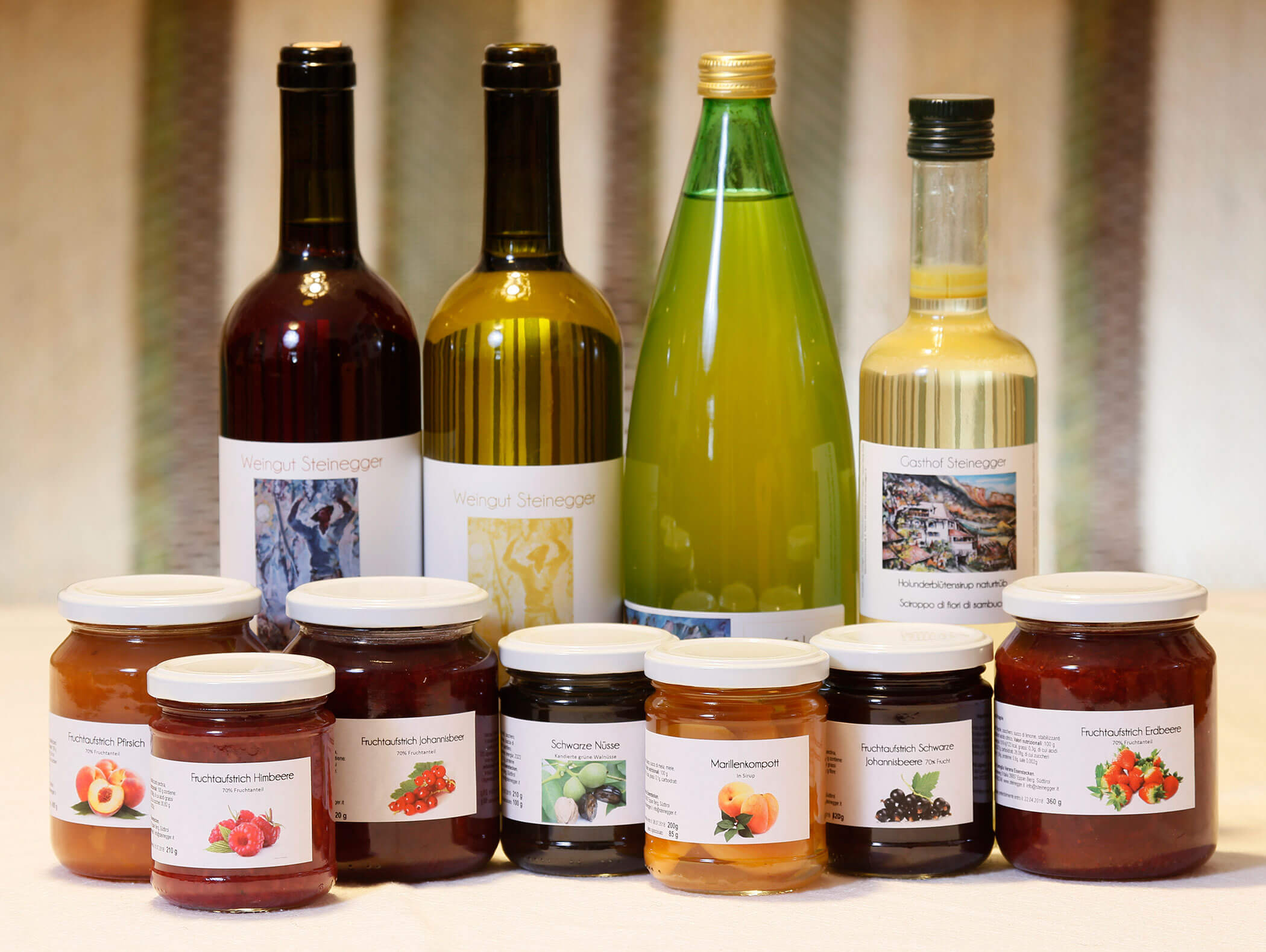 Bottiglie assortite di succo di frutta e vasetti di marmellata di frutta esposti su un tavolo con sfondo a righe. - Wandergasthof Steinegger