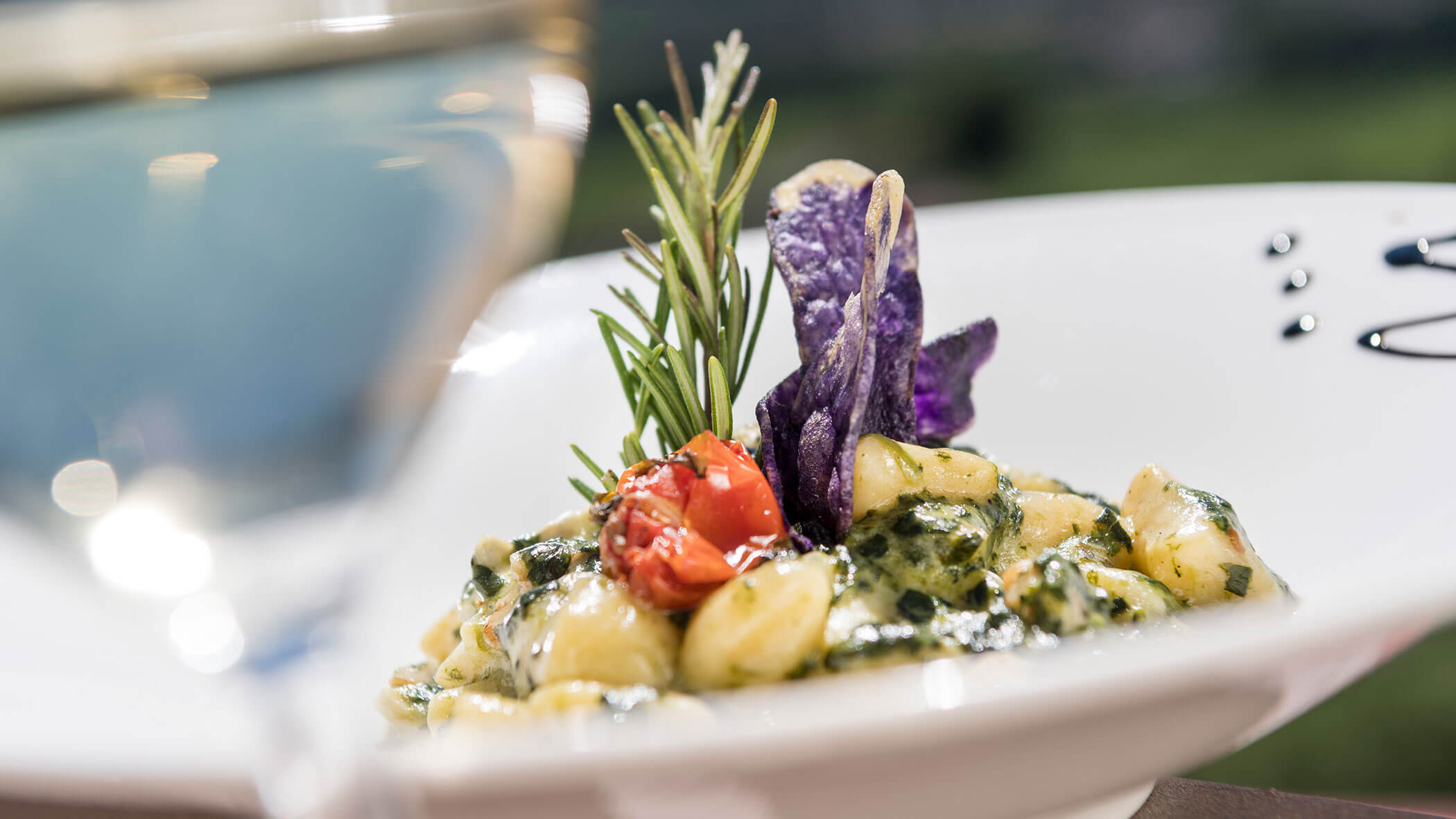 Eine Nahaufnahme von Gnocchi mit Pesto, Kirschtomaten, Kräutern und violetten Chips, neben einem Glas Weißwein. - Wandergasthof Steinegger