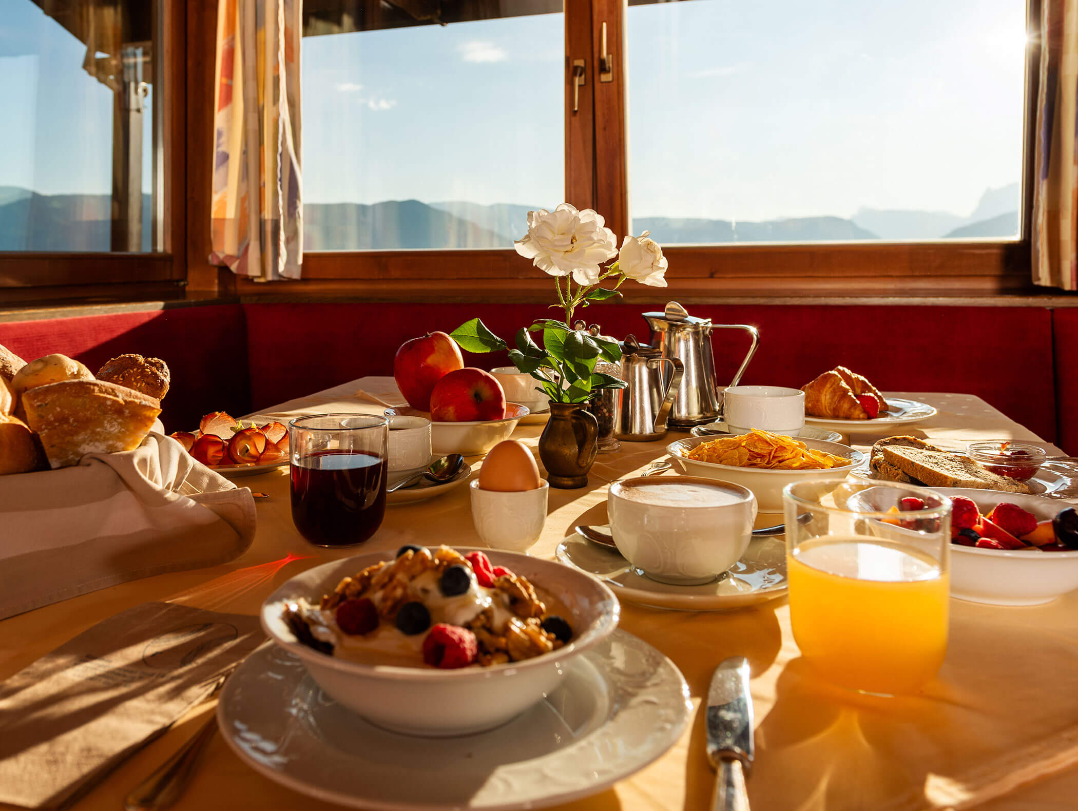 Ein sonnendurchfluteter Frühstückstisch mit Obst, Gebäck, Saft, Kaffee, Eiern und Blumen an einem Fenster mit Blick auf die Berge. - Wandergasthof Steinegger