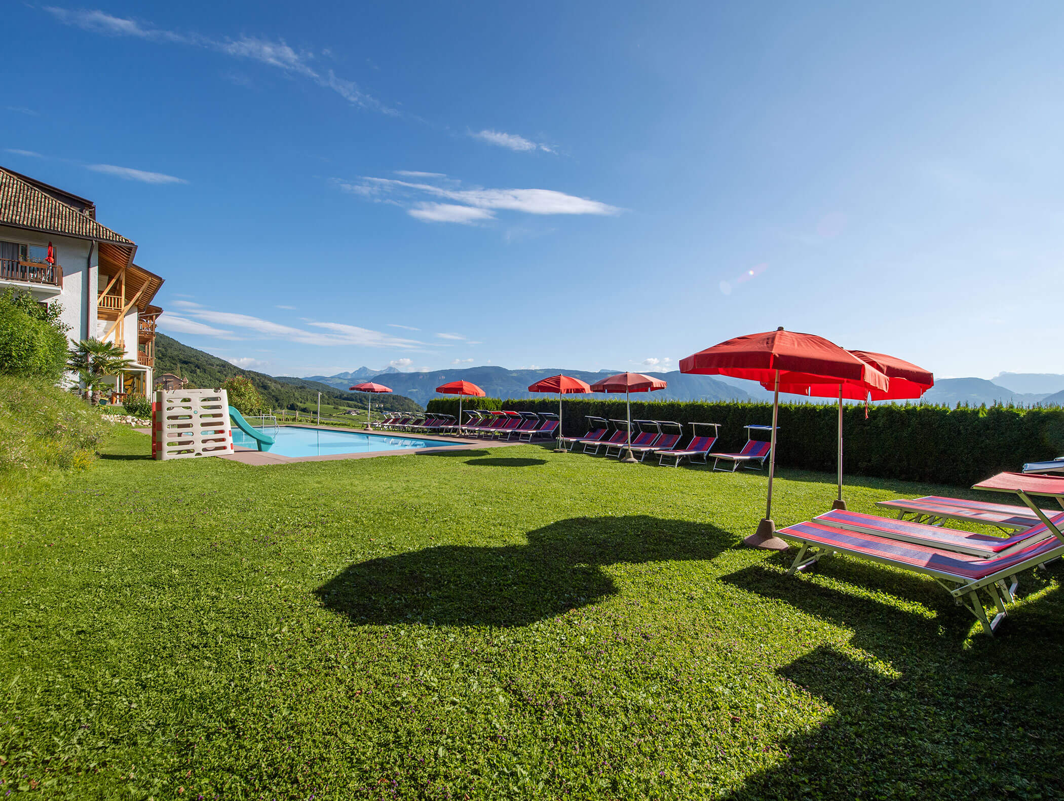 Lawn with lounge chairs and red umbrellas by a pool, mountains and clear sky in the background. - Wandergasthof Steinegger
