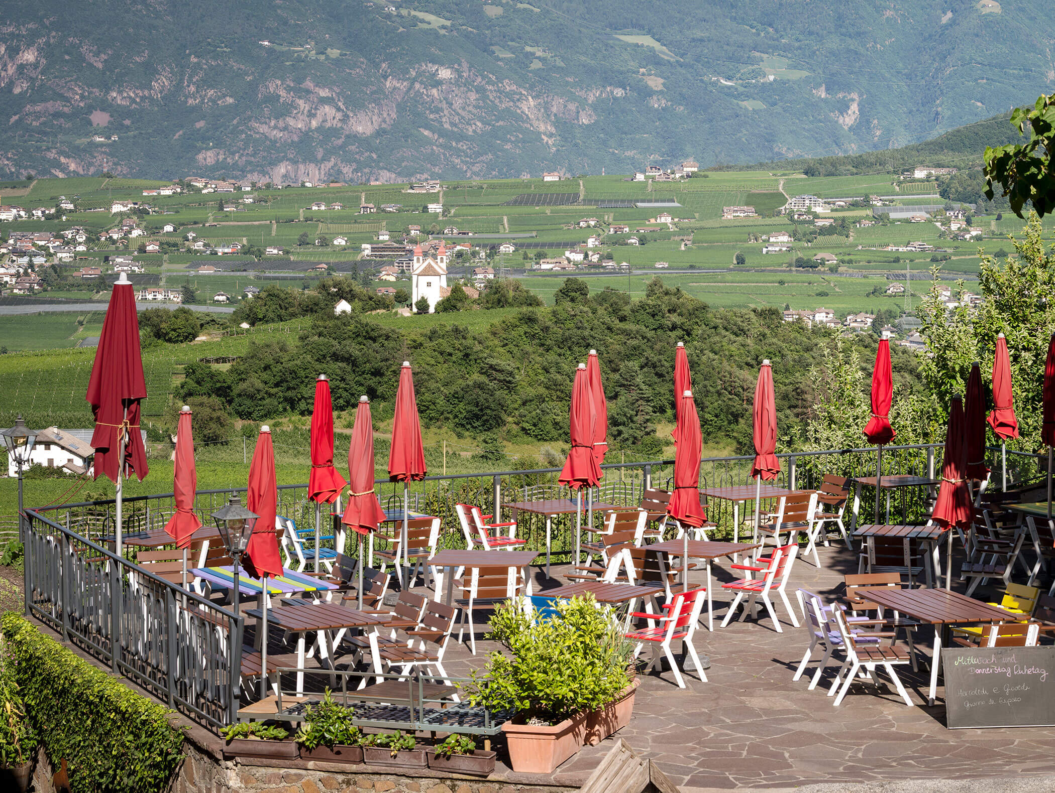Outdoor café with red umbrellas and empty tables, overlooking a valley with vineyards and distant mountains. - Wandergasthof Steinegger