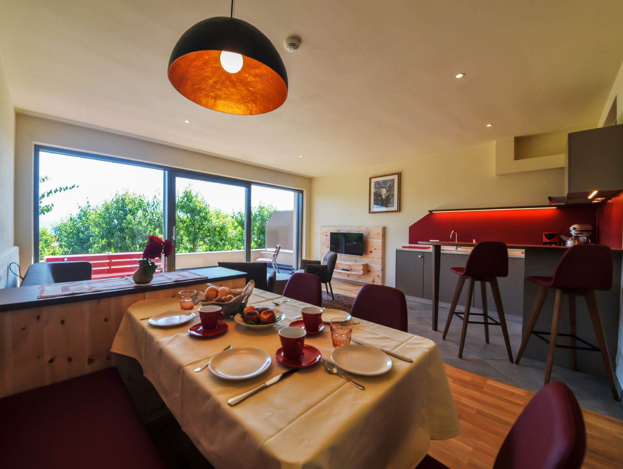 Modern kitchen and dining area with large windows, a set table, and red accents in natural sunlight. - Wandergasthof Steinegger