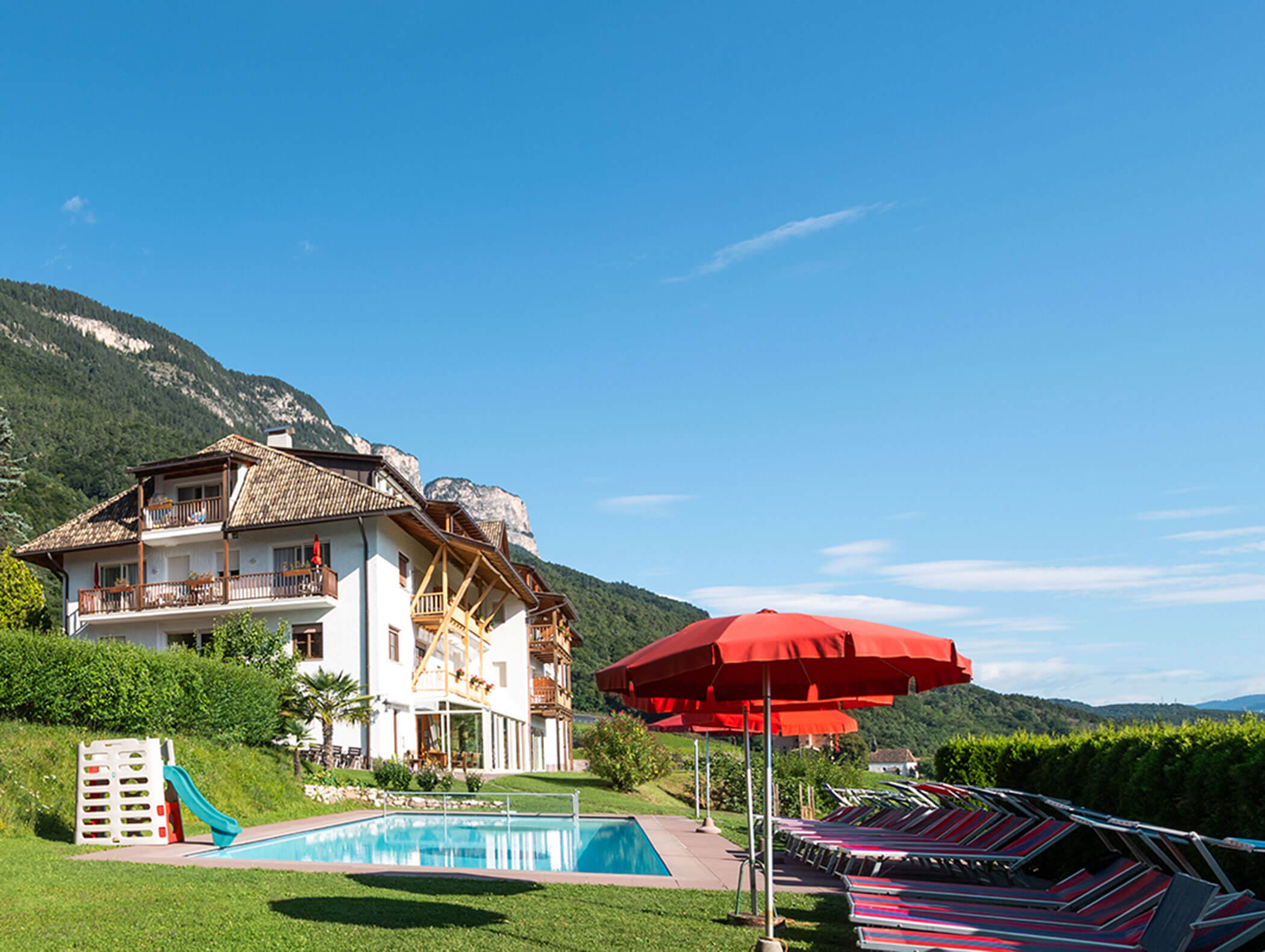 Hotel con balconi a bordo piscina, ombrelloni rossi, lettini e montagne sotto un cielo azzurro e limpido. - Wandergasthof Steinegger