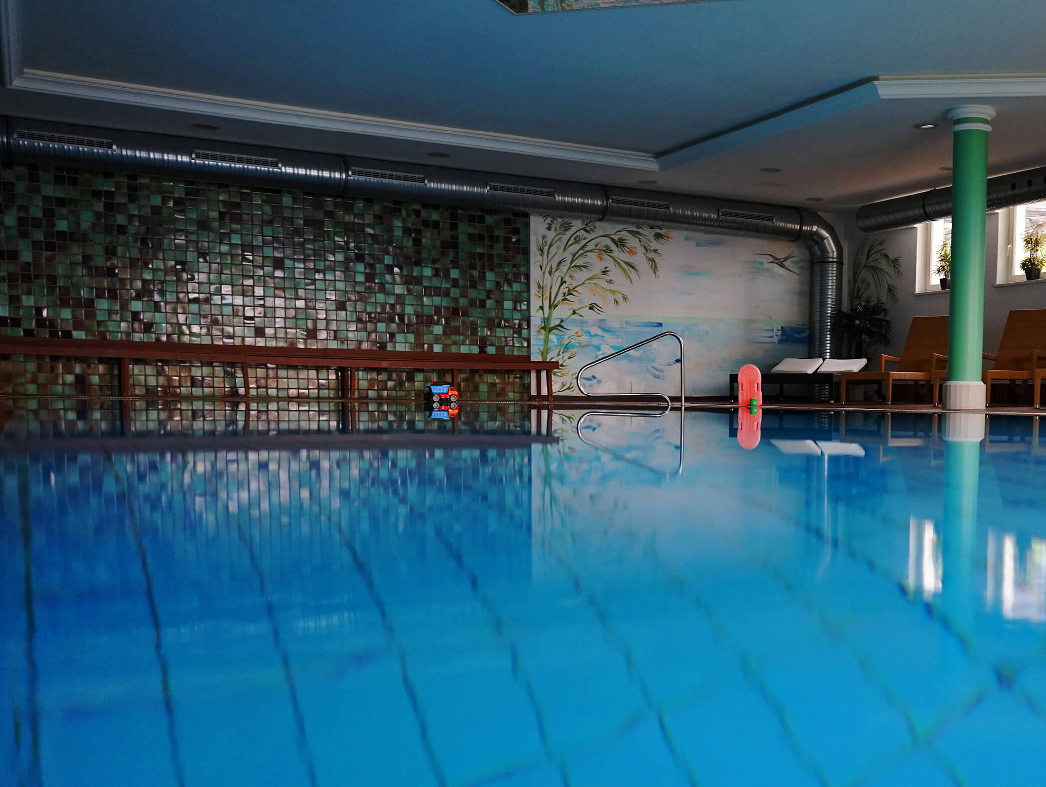 Piscina coperta con acqua blu, pareti piastrellate, panchine e un murale dipinto sullo sfondo. - Wandergasthof Steinegger