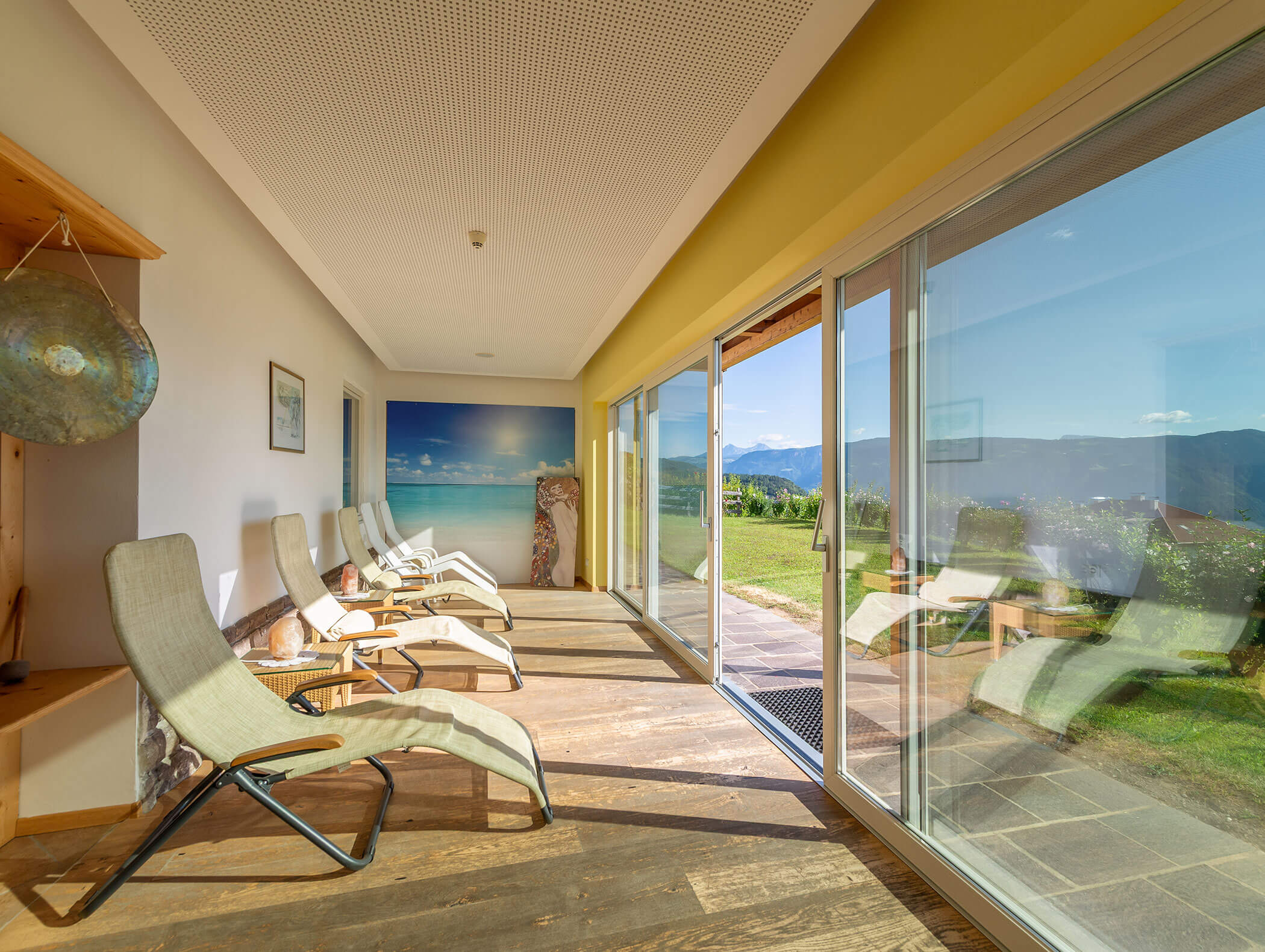 Sala relax illuminata dal sole con sedie a sdraio, ampie finestre e una vista panoramica sulle colline verdi e il cielo blu all'esterno. - Wandergasthof Steinegger