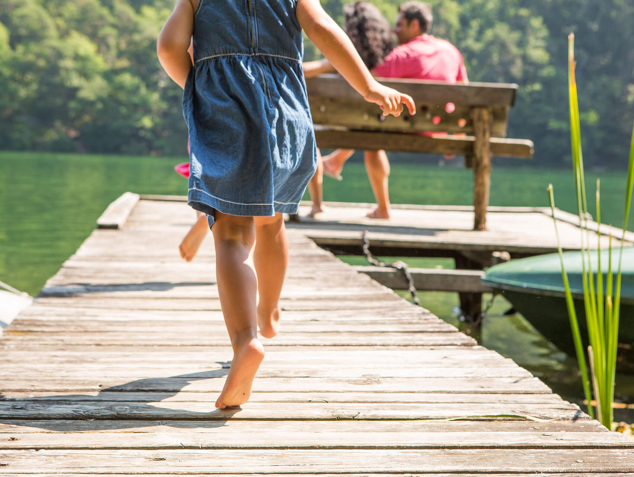 Una bambina con un vestito di jeans cammina a piedi nudi su una banchina di legno verso una famiglia seduta in riva al lago. - Wandergasthof Steinegger