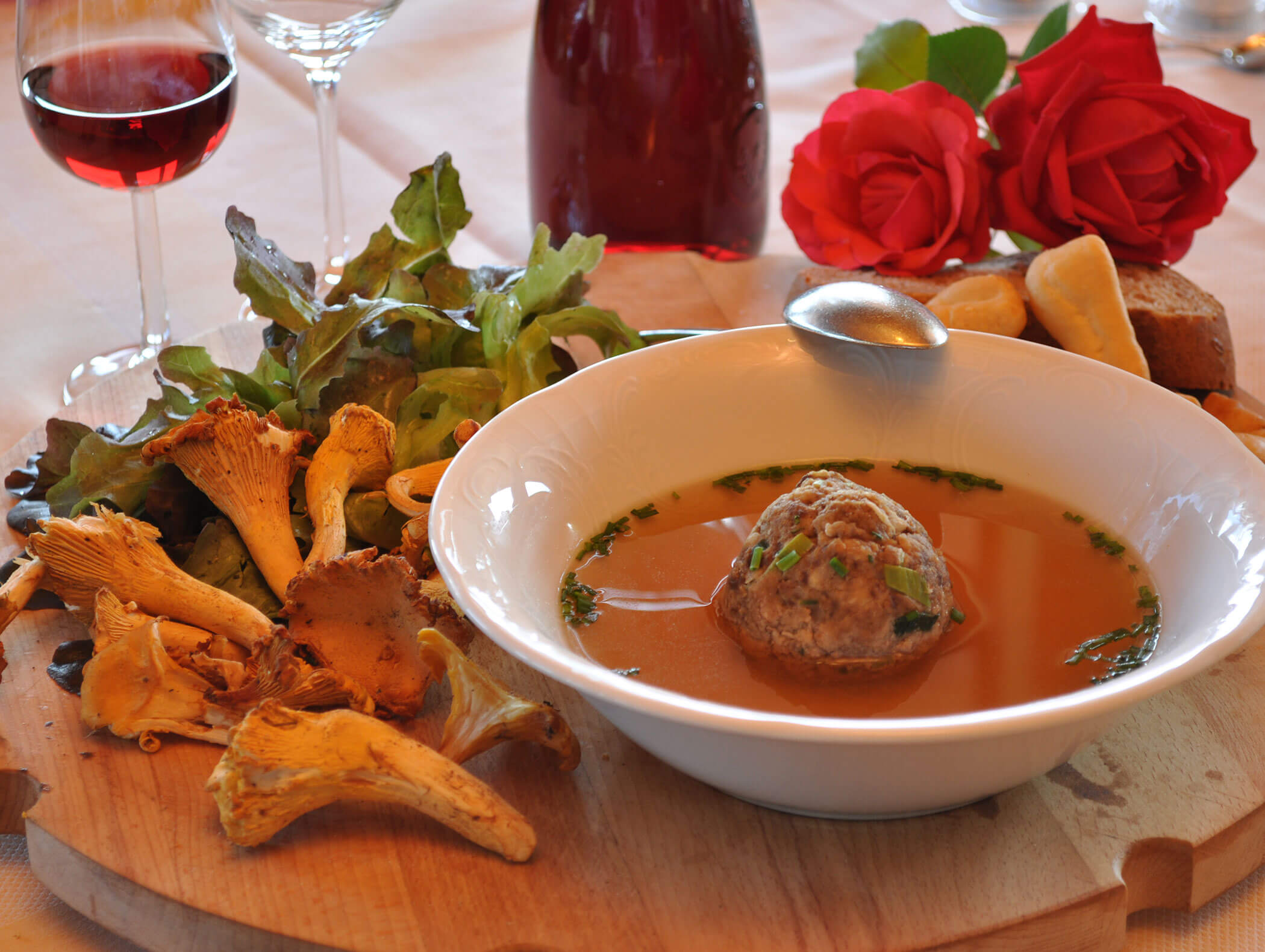 Ciotola di zuppa con gnocco, verdure fresche, funghi, pane e rose su una tavola di legno. - Wandergasthof Steinegger