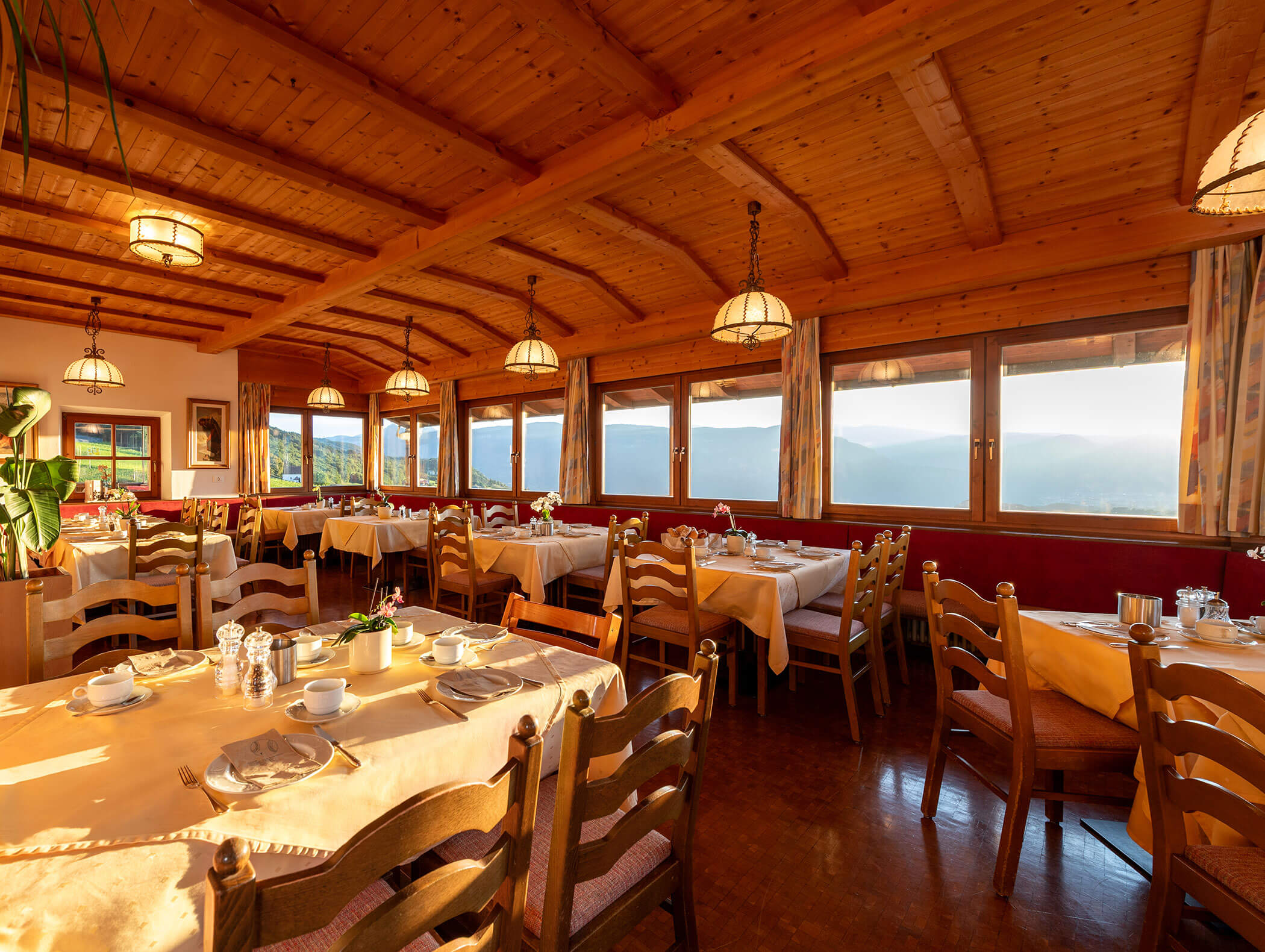 Sala da pranzo illuminata dal sole con soffitto in legno, tavoli apparecchiati e vista sulle montagne attraverso grandi finestre. - Wandergasthof Steinegger