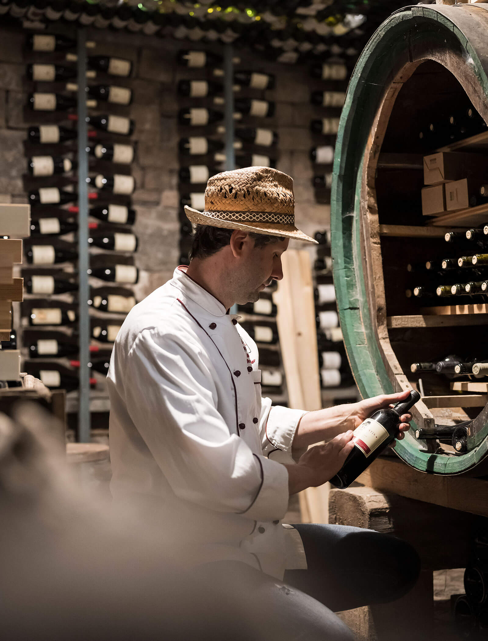 Uno chef con un cappello di paglia esamina una bottiglia di vino in una cantina poco illuminata, circondata da scaffali di bottiglie. - Wandergasthof Steinegger