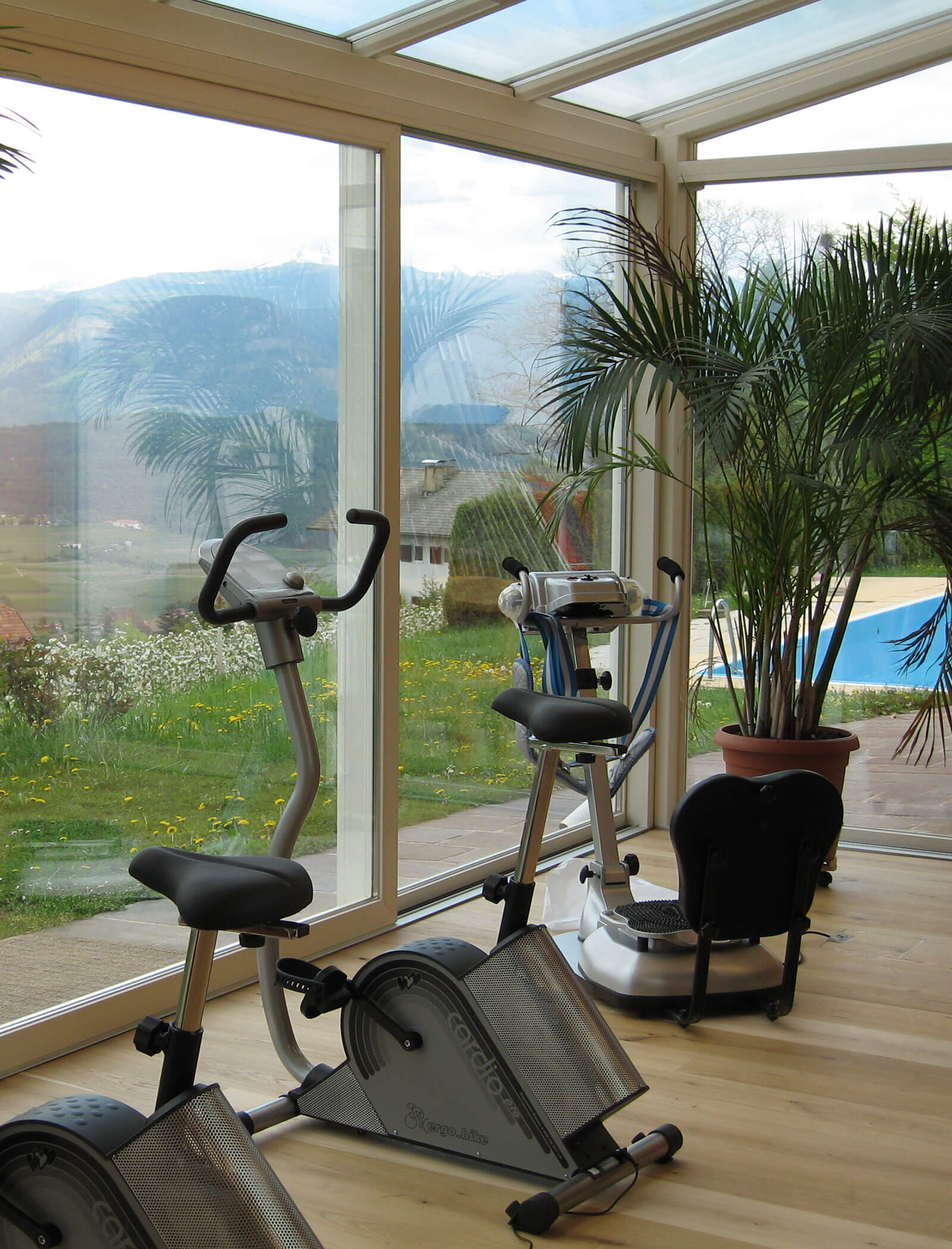 Two exercise bikes in a sunlit room with large windows overlooking a scenic outdoor landscape and greenery. - Wandergasthof Steinegger