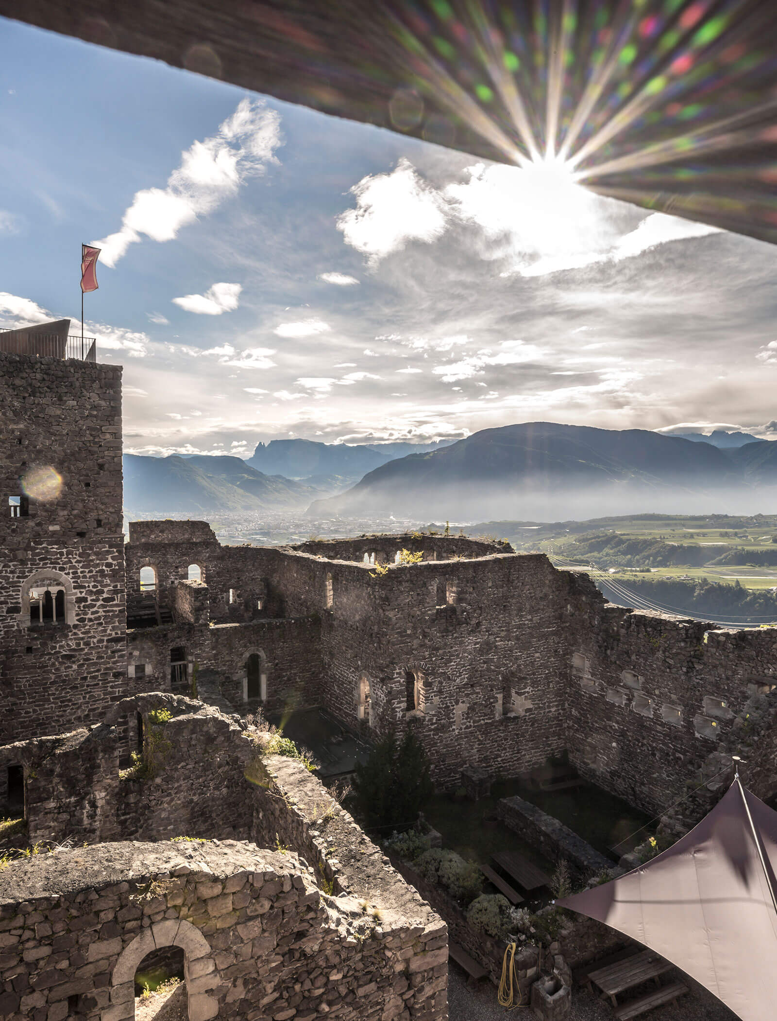 Sunlight bursts over a stone castle ruin with mountains and a valley in the background. - Wandergasthof Steinegger