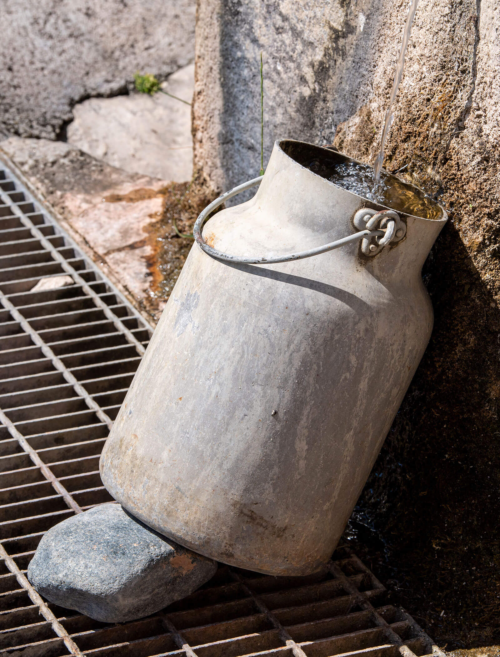 Eine Milchkanne aus Metall, die über einen Ausguss mit Wasser gefüllt wird und mit einem Stein auf ein Gitter gestützt ist. - Wandergasthof Steinegger