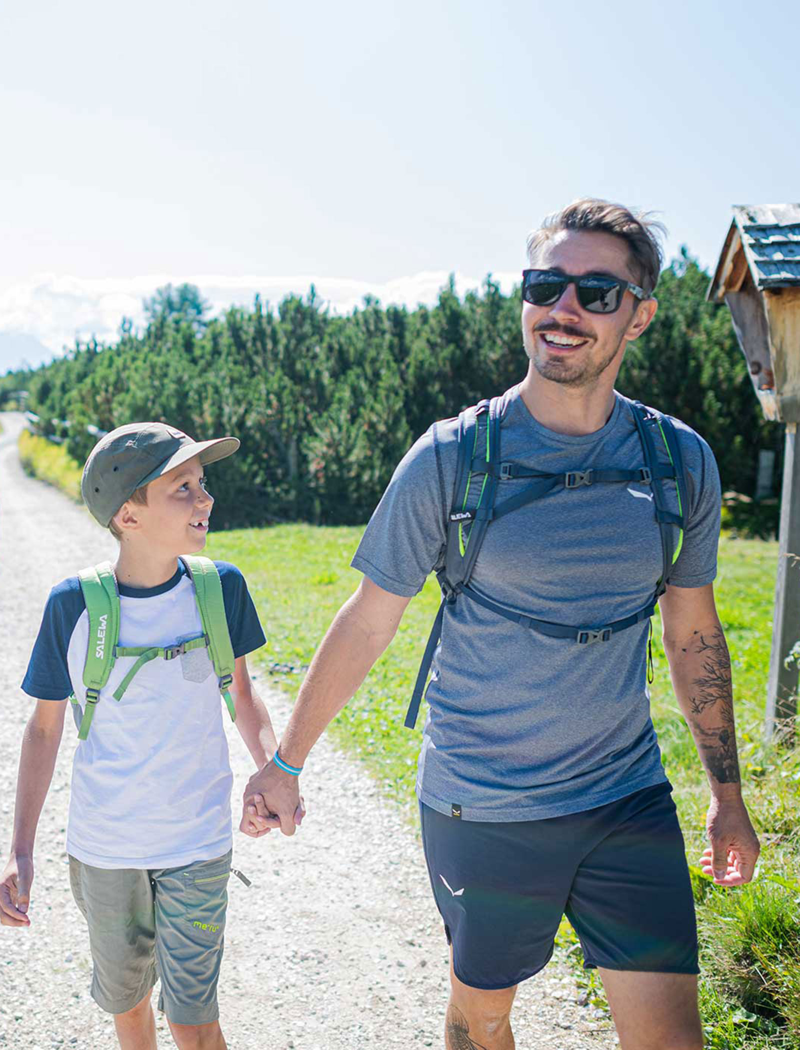 A man and boy, both with backpacks, smile and hold hands while hiking on a sunny trail. - Wandergasthof Steinegger