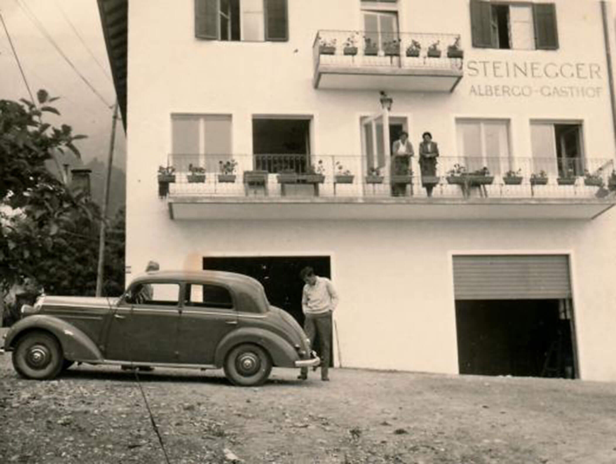 A man stands by a vintage car in front of a white building with balconies and STEINEGGER signage. - Wandergasthof Steinegger