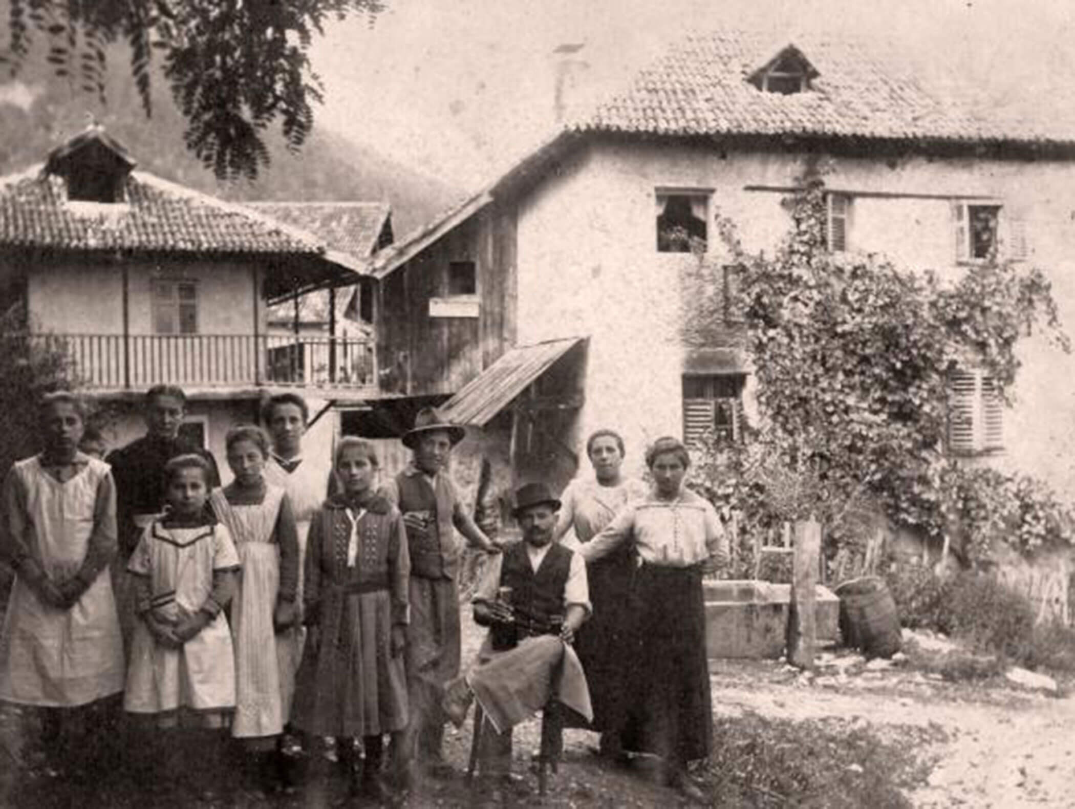 A group of people in old-fashioned clothing pose in front of rustic houses covered in vines. - Wandergasthof Steinegger