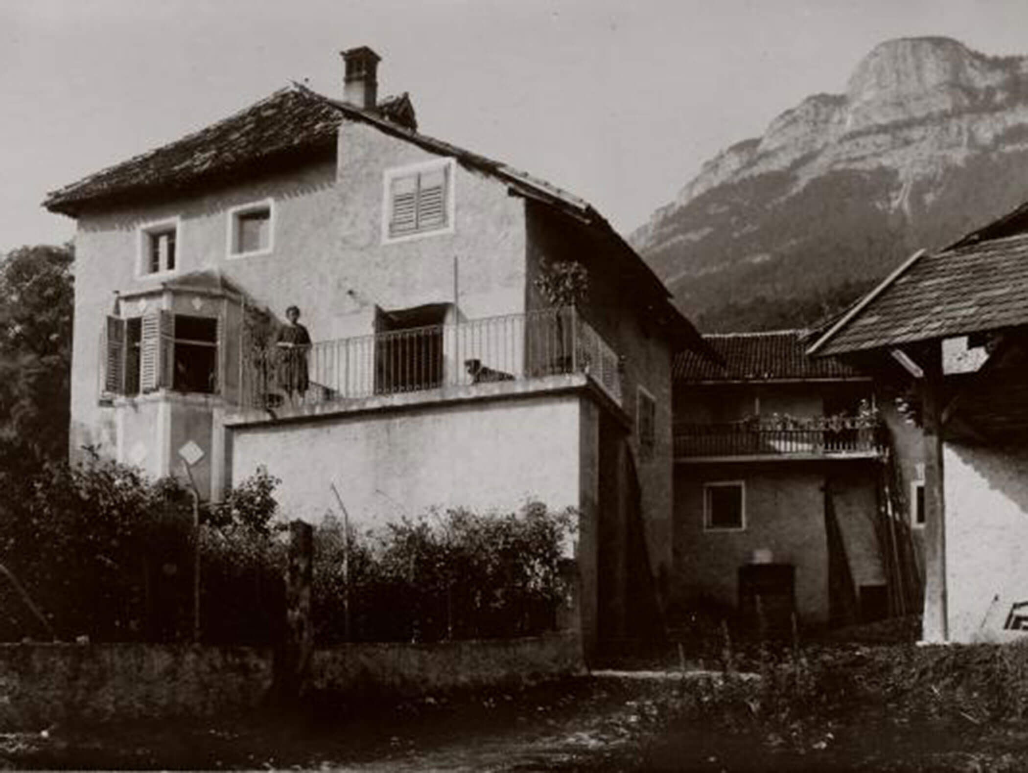 Old house with a balcony, a person standing on it, mountains in the background, and shrubs in the foreground. - Wandergasthof Steinegger