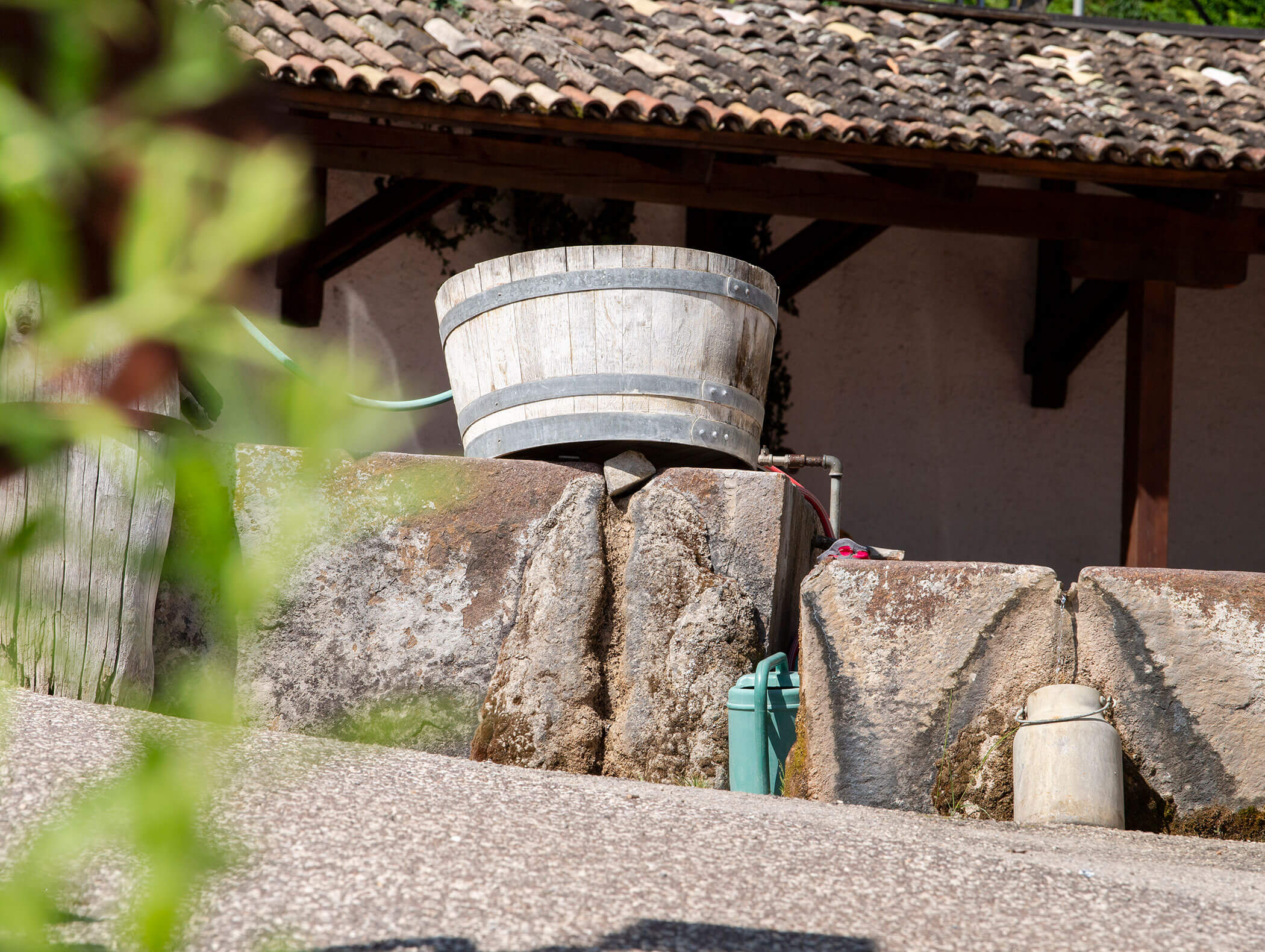 A wooden barrel sits on stone slabs near a tiled roof, with a green hose and jug nearby. - Wandergasthof Steinegger