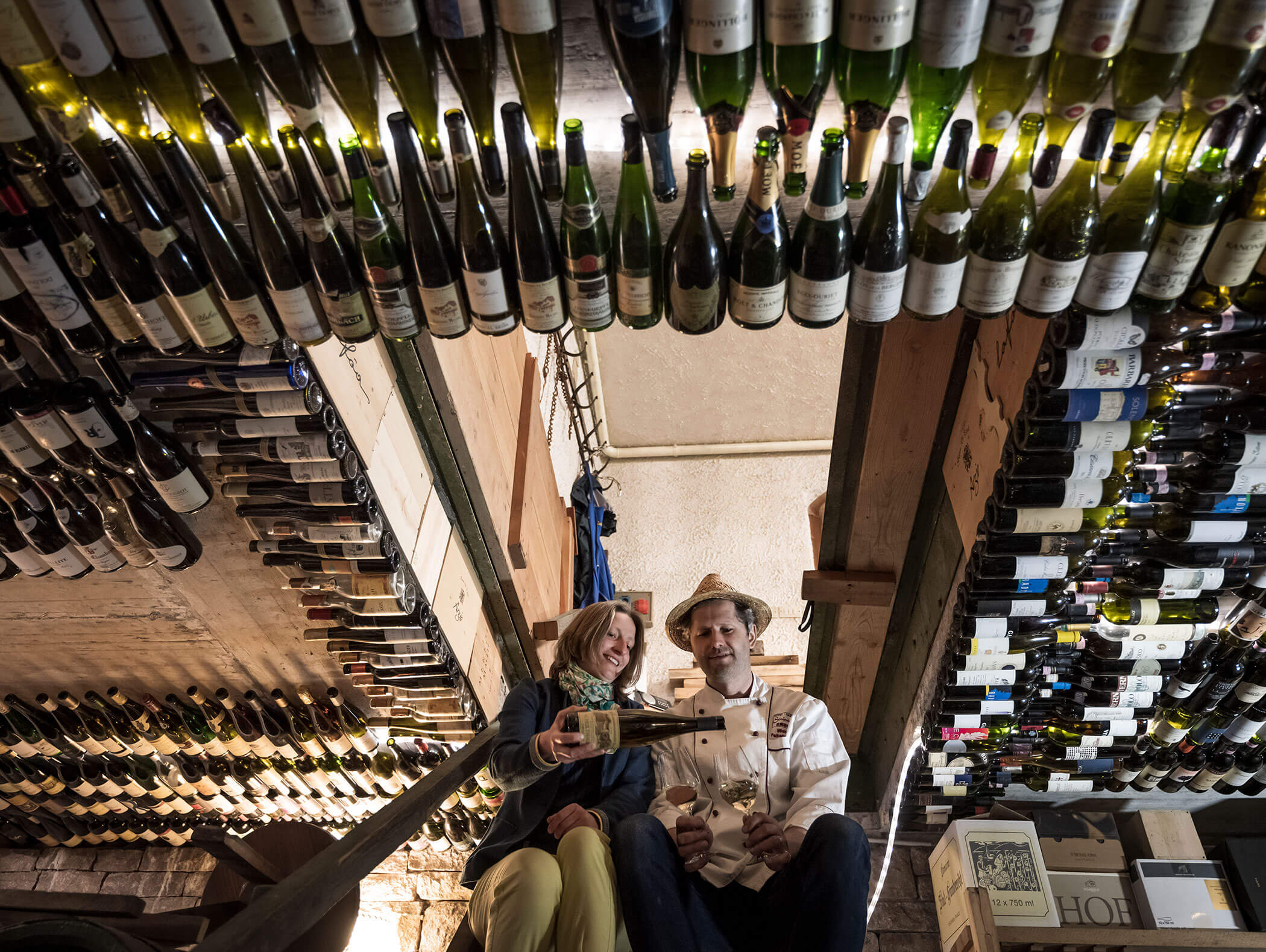 Two people sit in a wine cellar, surrounded by shelves and rows of wine bottles, holding glasses and a bottle. - Wandergasthof Steinegger