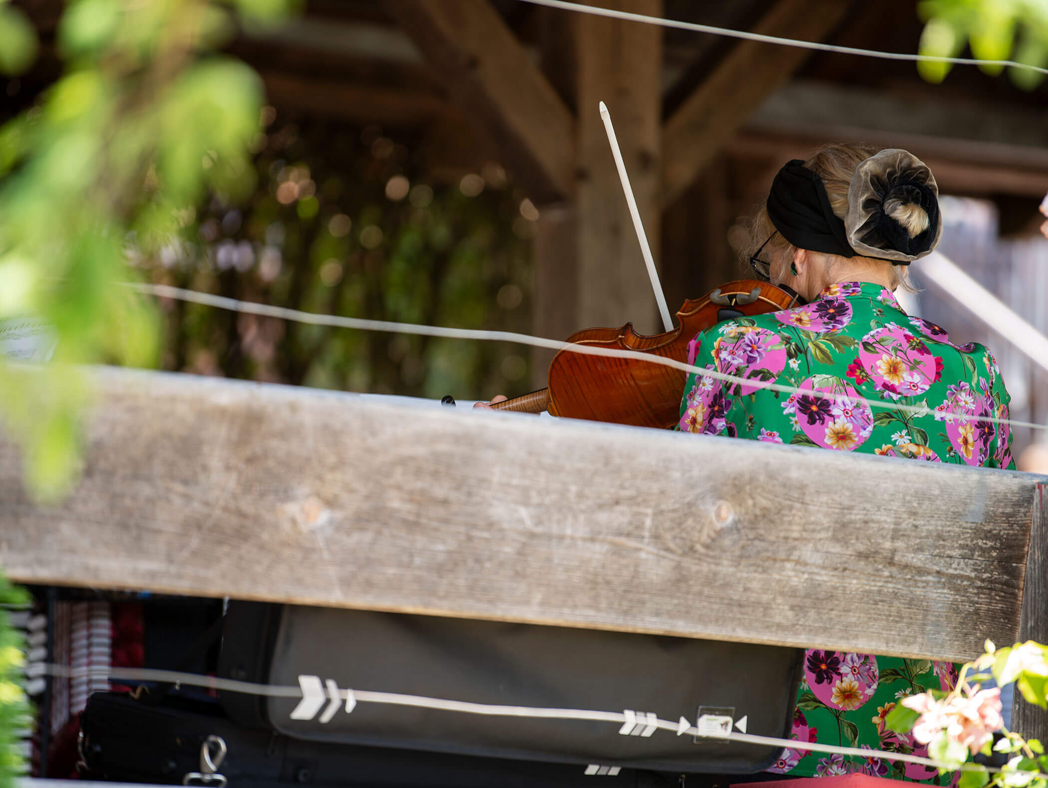 Woman in a floral dress playing a violin outdoors, viewed from behind through a wooden railing. - Wandergasthof Steinegger