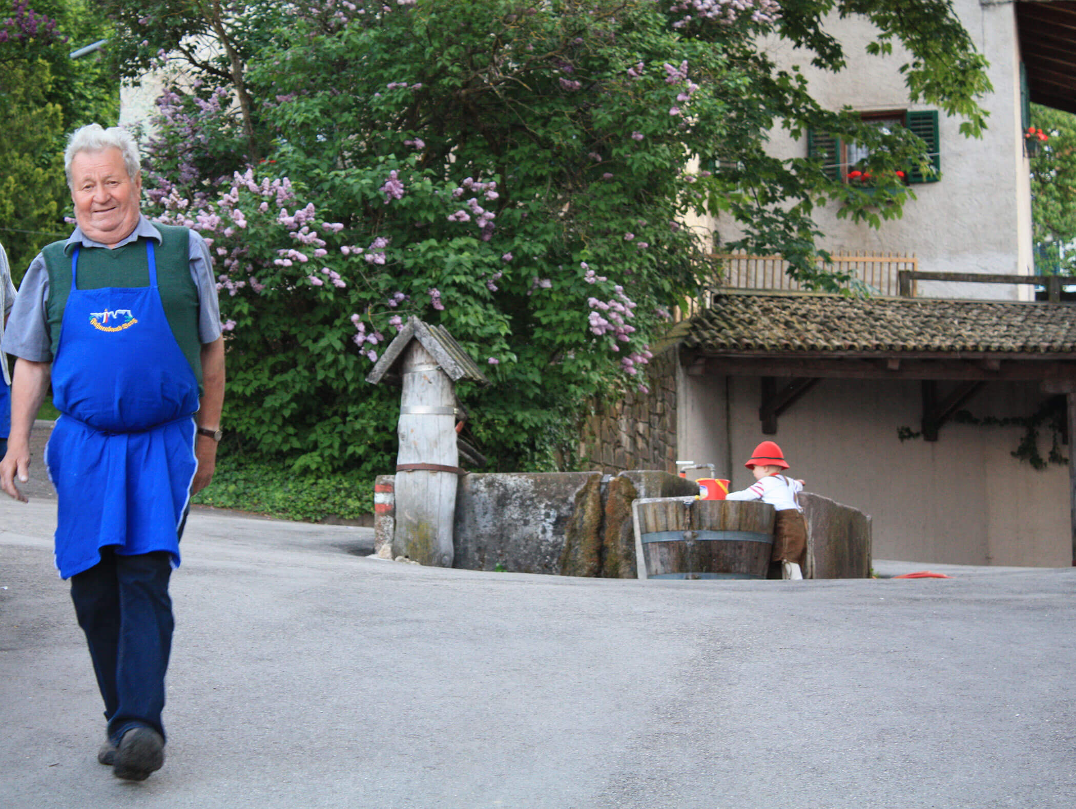 Smiling man in blue apron walks on a street near a rustic house and blooming bushes on a sunny day. - Wandergasthof Steinegger