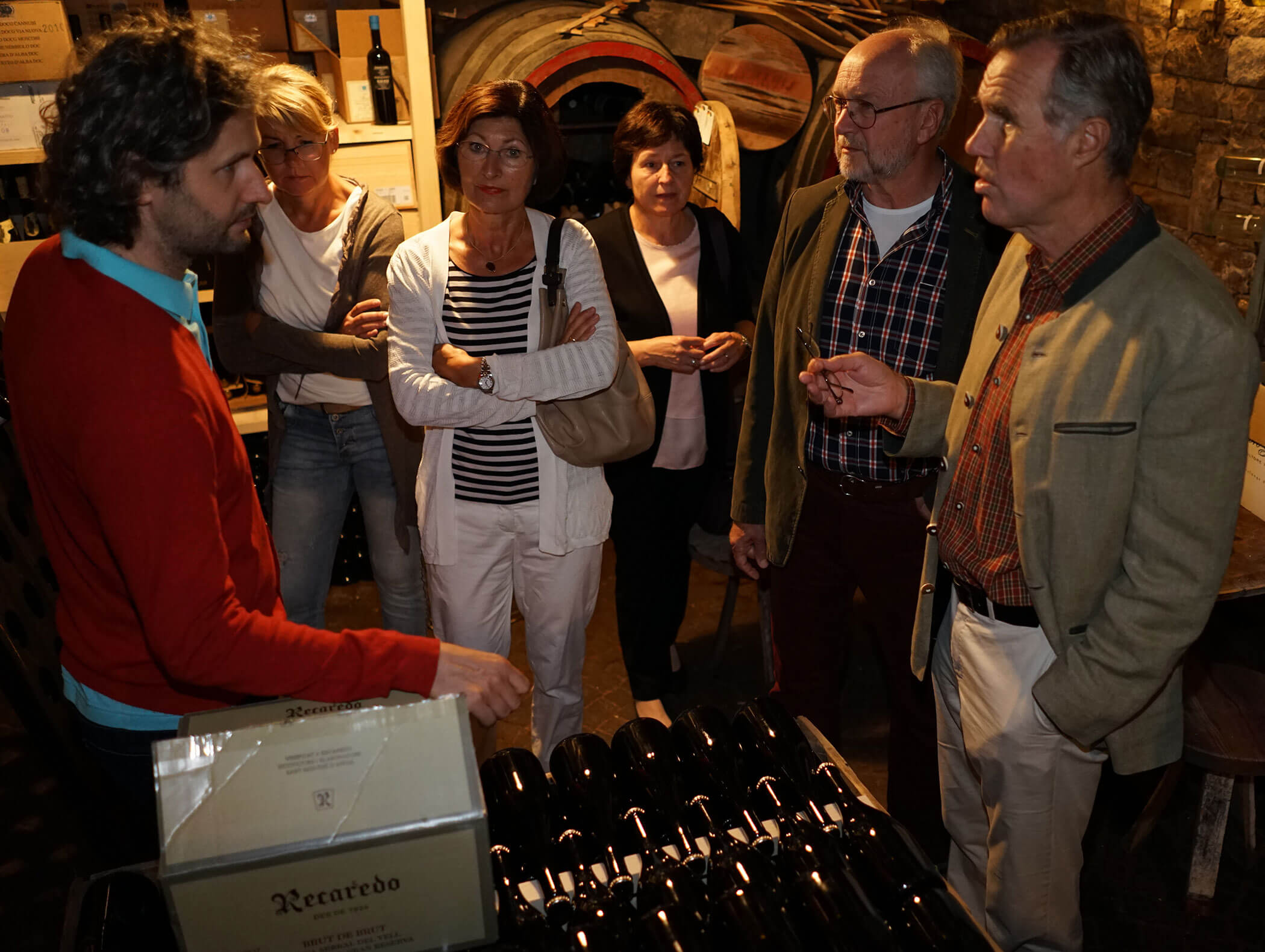 Six adults stand in a wine cellar, listening to a man explaining near racks of wine bottles. - Wandergasthof Steinegger