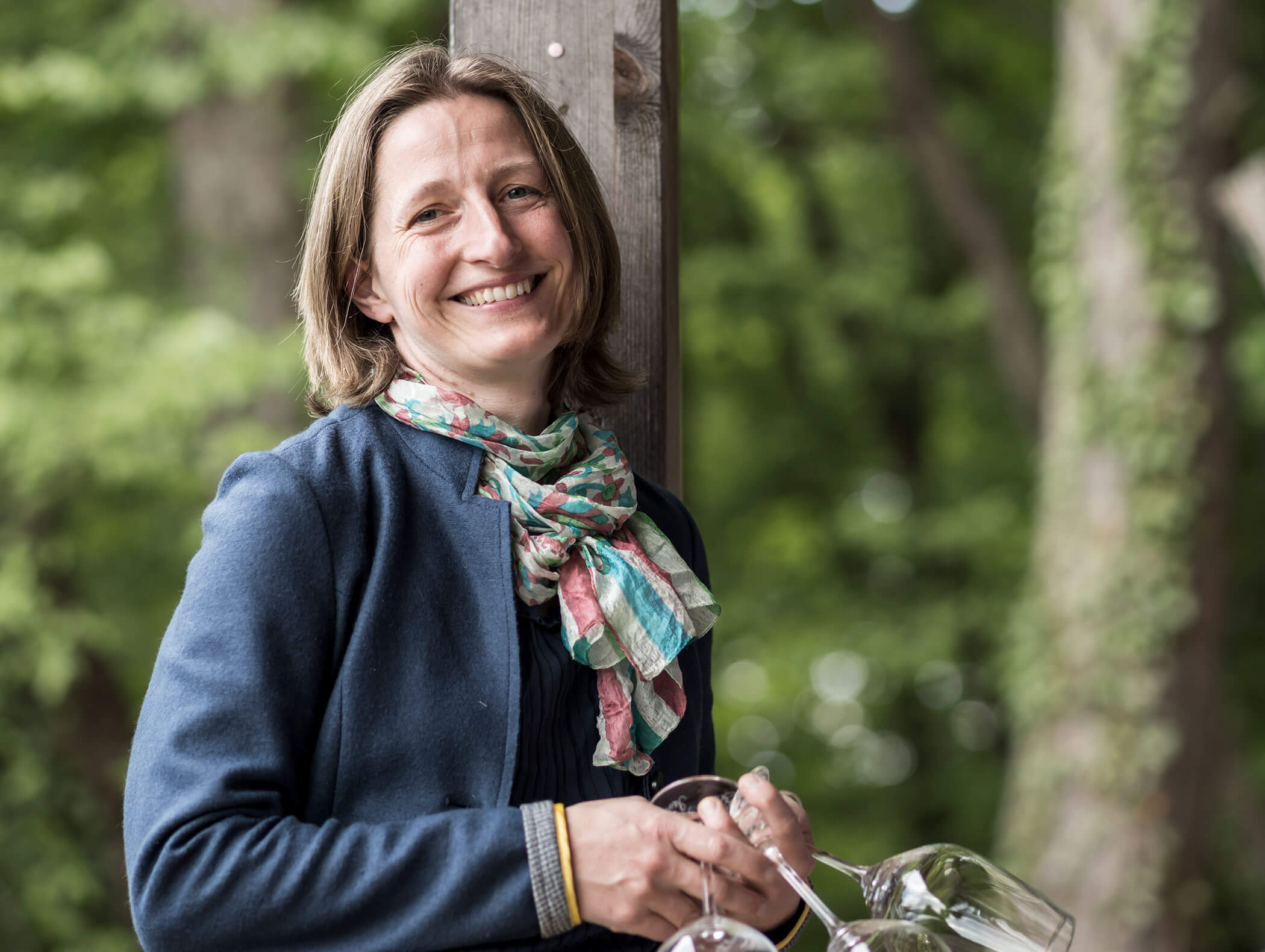 Smiling woman in a blue jacket and scarf holding empty wine glasses outdoors with trees in the background. - Wandergasthof Steinegger