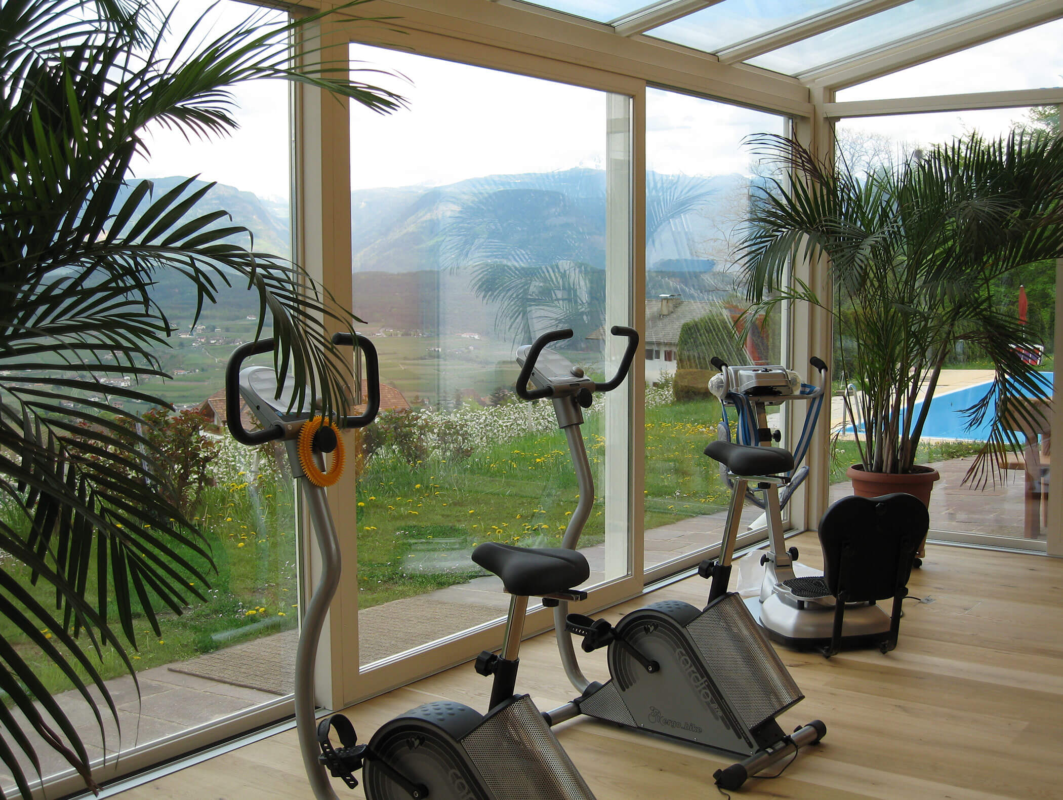 Zwei Heimtrainer in einem hellen Raum mit großen Fenstern, mit Blick auf eine malerische Berglandschaft und Grünflächen. - Wandergasthof Steinegger