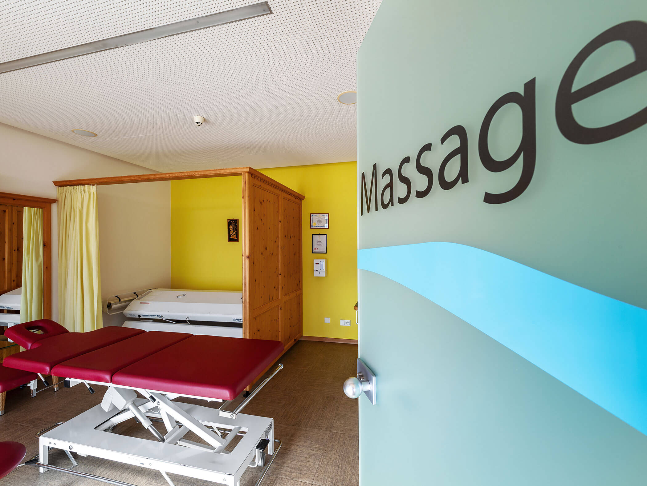 Massageraum mit zwei roten Massagetischen, einem Bett, gelben Wänden und einer teilweise geöffneten Tür mit der Aufschrift Massage. - Wandergasthof Steinegger