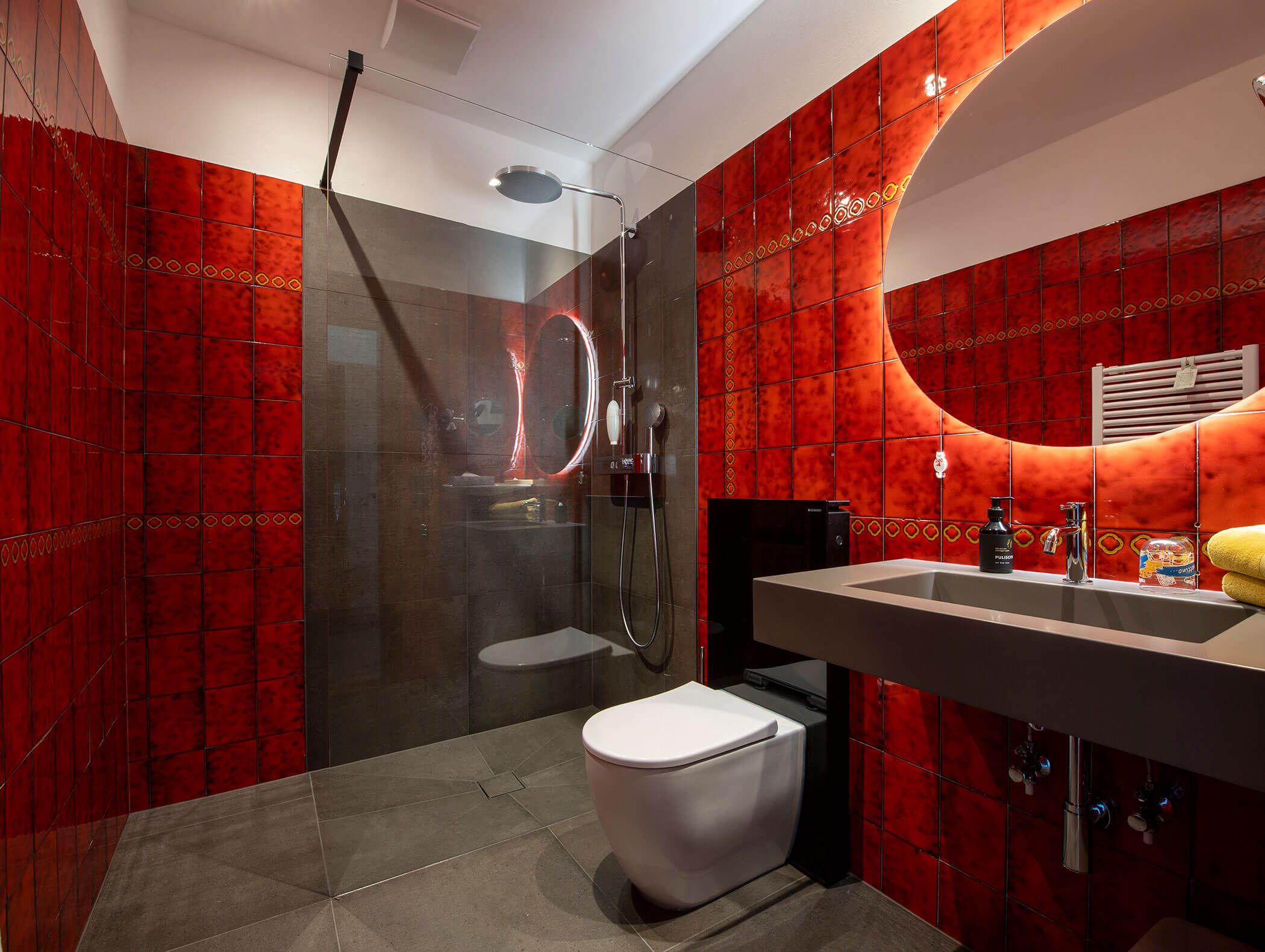 Modern bathroom with red tiled walls, glass shower, wall-mounted toilet, and large illuminated round mirror. - Wandergasthof Steinegger