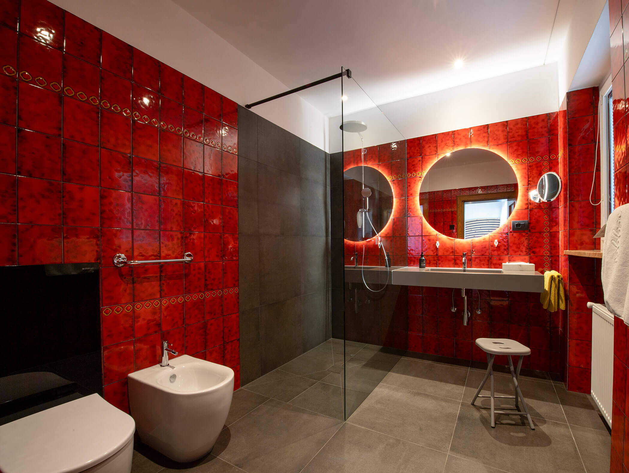 Modern bathroom with red tiled walls, large mirrors, walk-in shower, bidet, and gray tile flooring. - Wandergasthof Steinegger